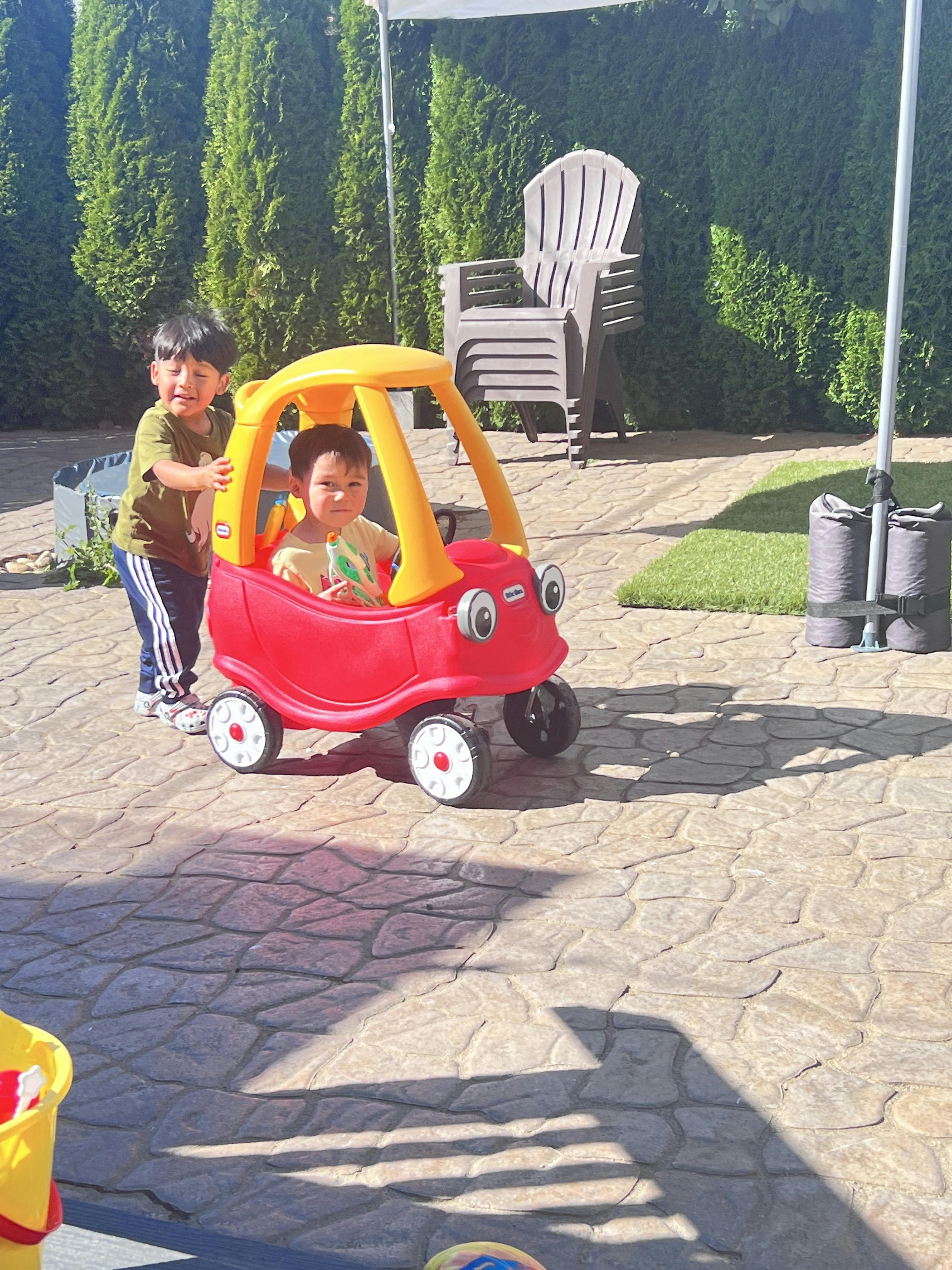 Child pushing another child in a red and yellow toy car outdoors