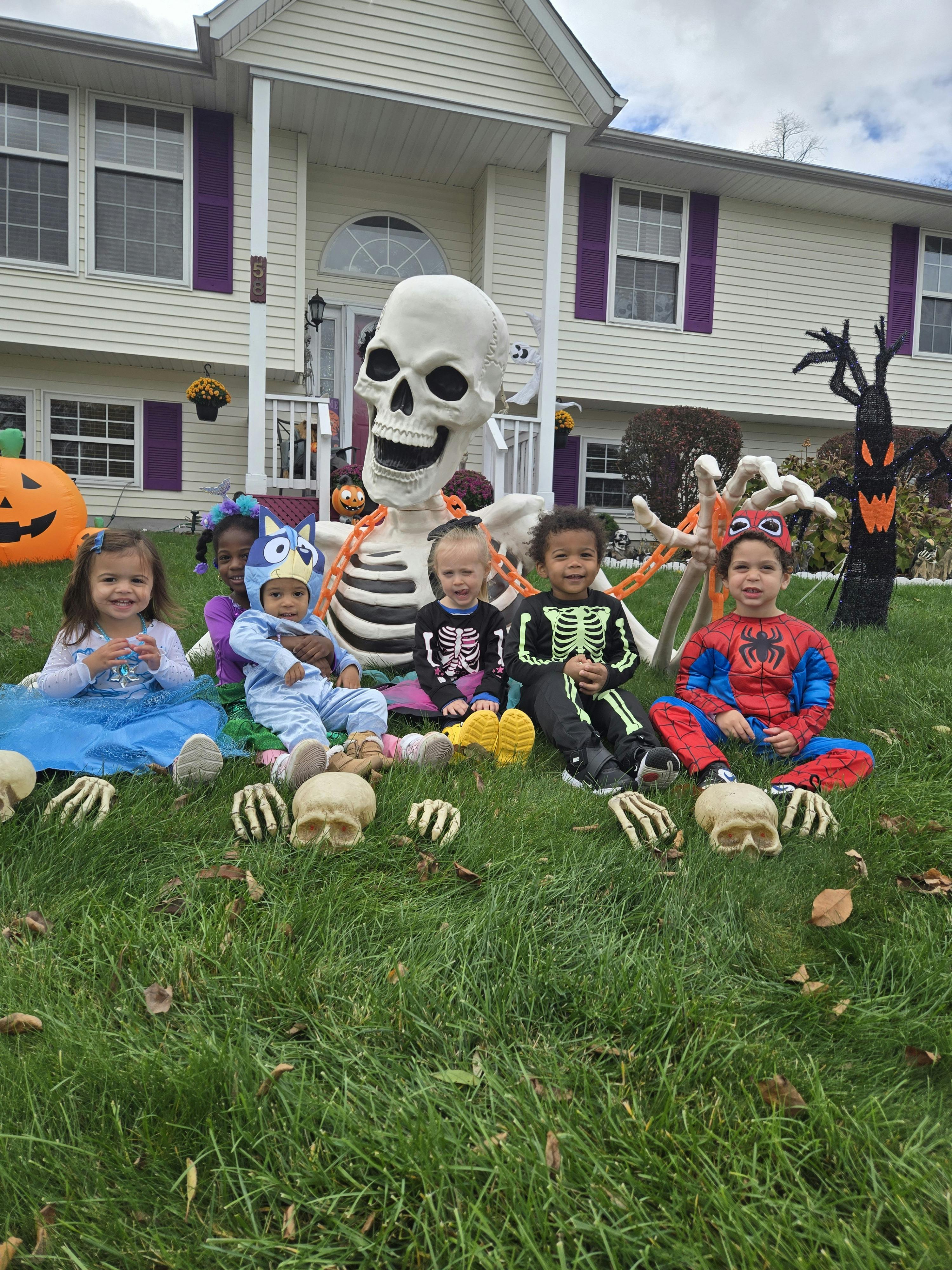 Five children in Halloween costumes sitting in front of a giant skeleton decoration
