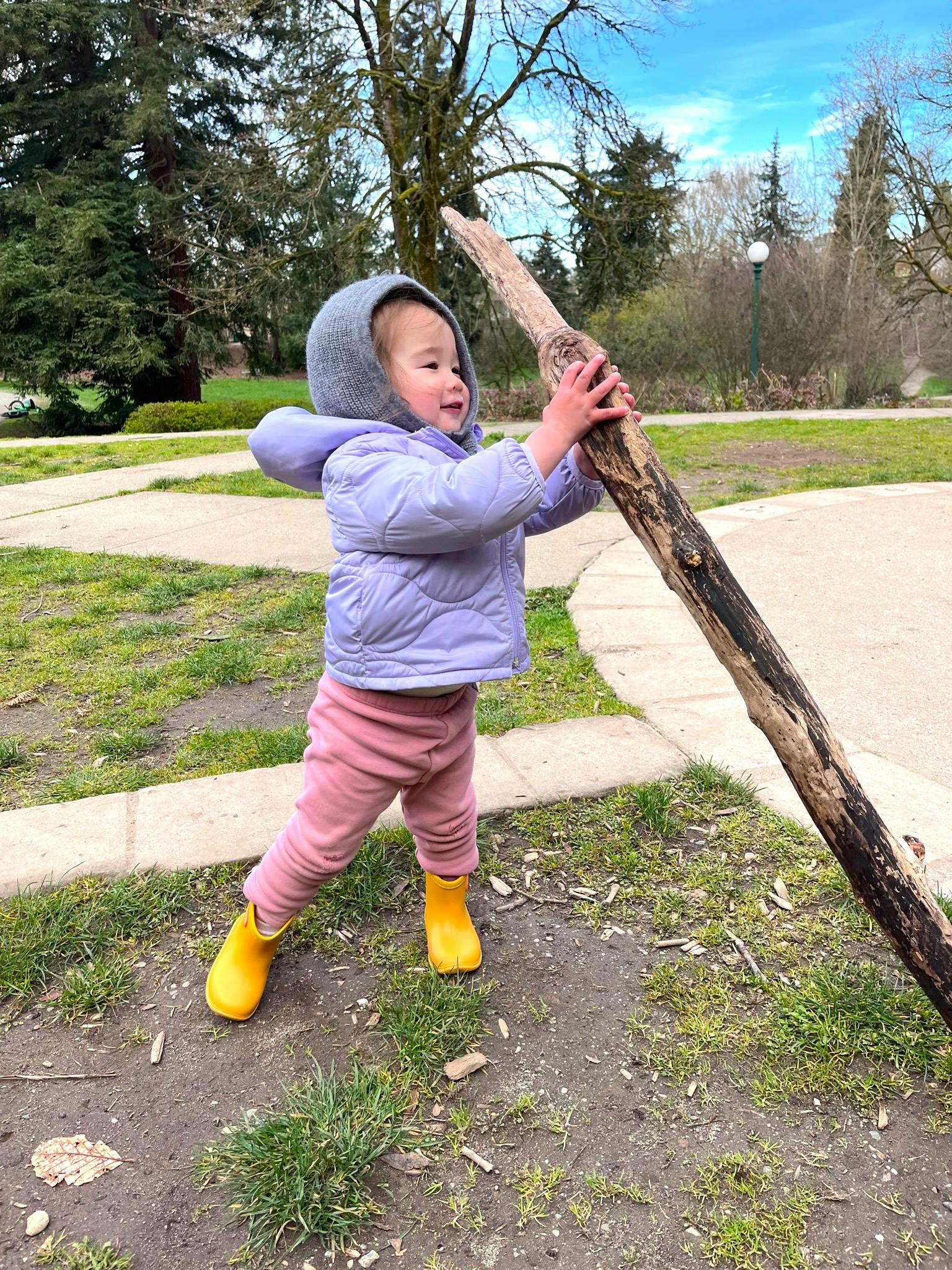 Child in a purple coat and yellow boots holding a large stick in a park