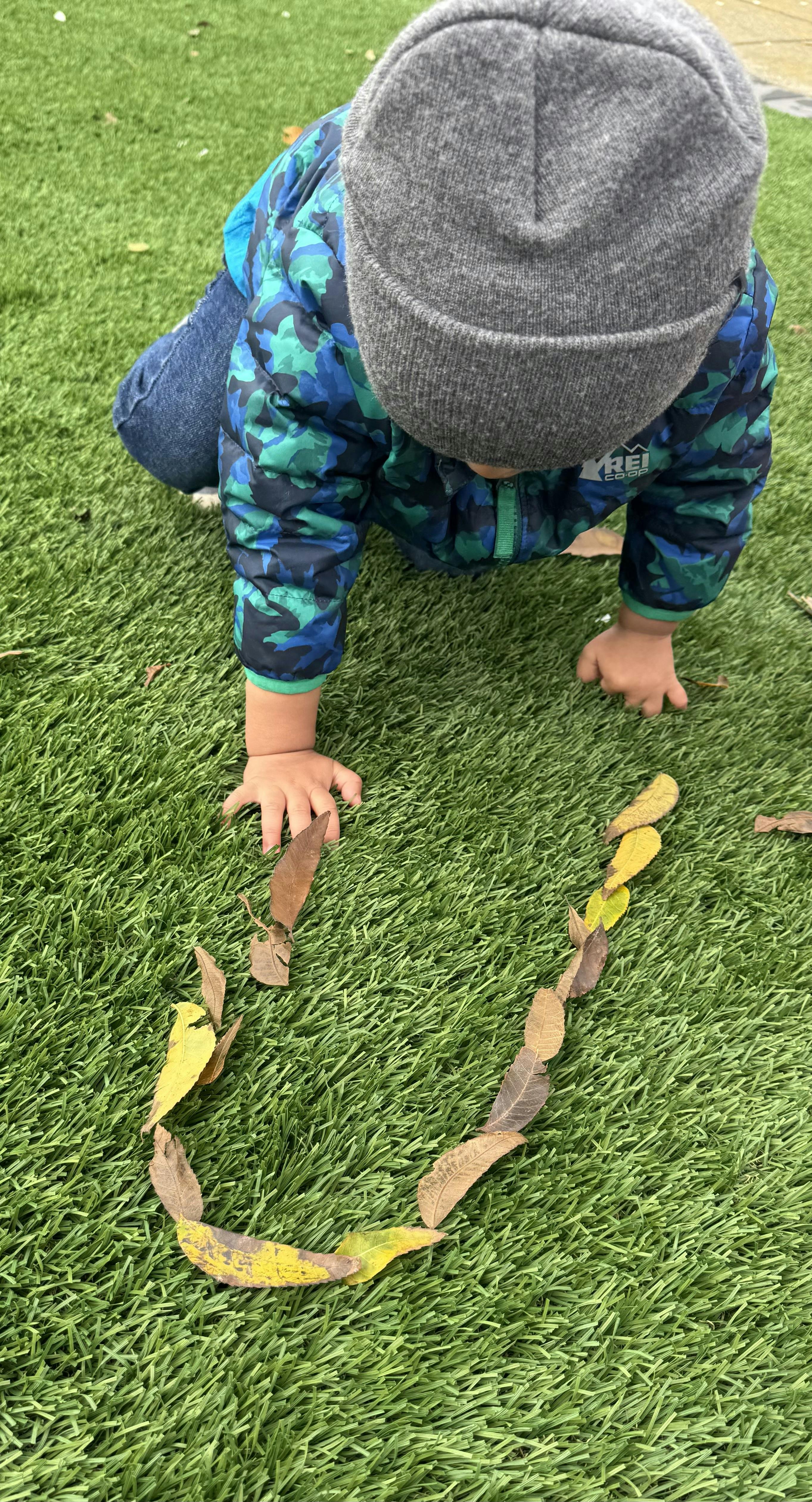 Child in a hat and jacket crawling on green grass near a line of leaves