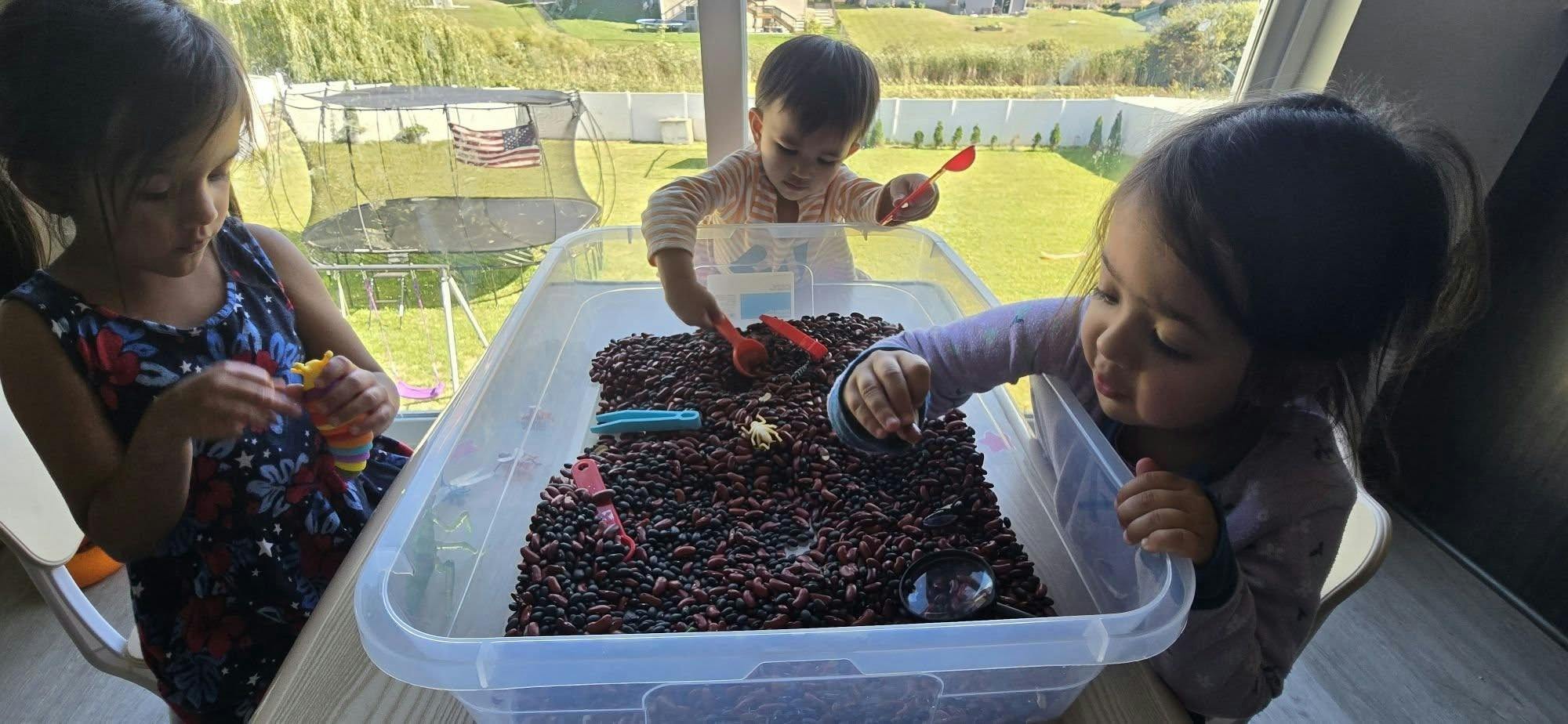 Three children playing with dried beans in a plastic storage bin