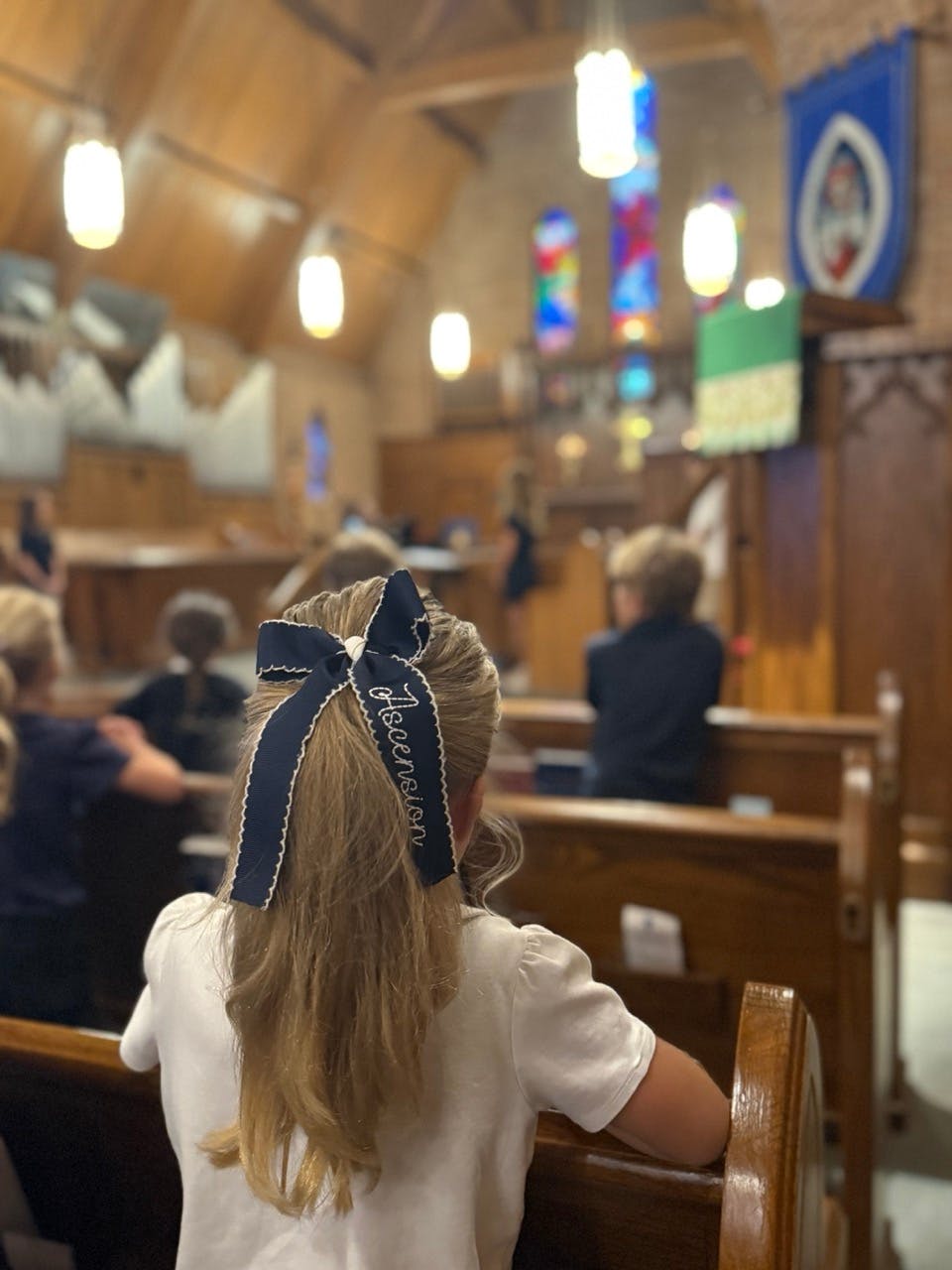 Child with an "Ascension" bow in their hair, seated in church pew