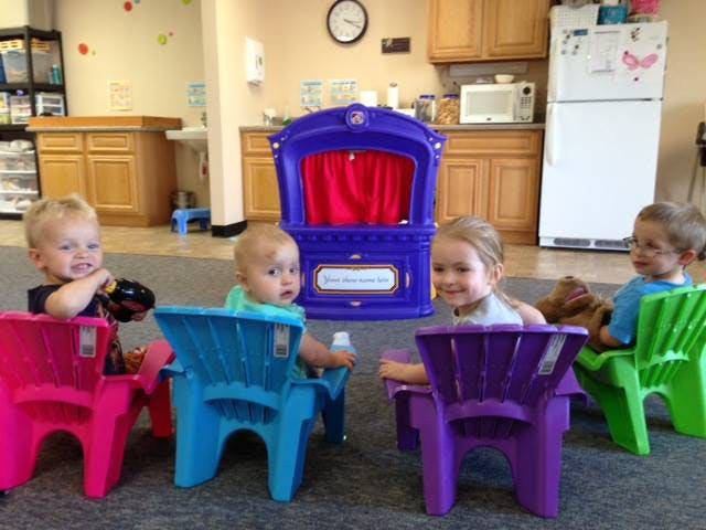 Four children sitting in colorful chairs in a room with a puppet theater