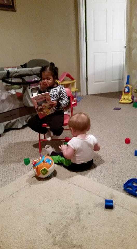 Child sitting on a chair reading a book next to a child on the floor