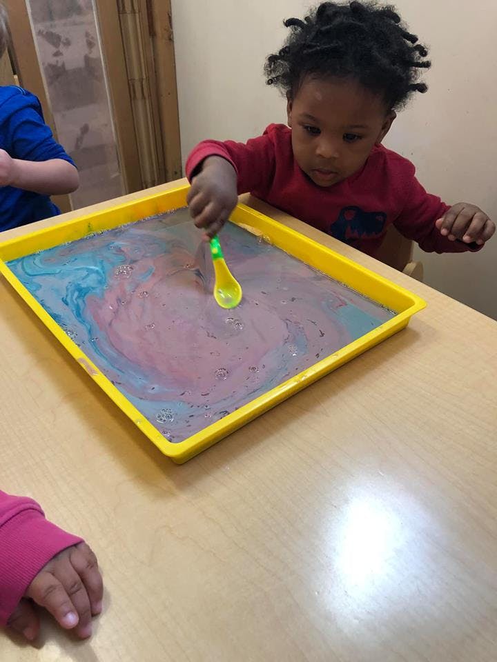 Child stirring colorful liquid in a yellow tray with a spoon