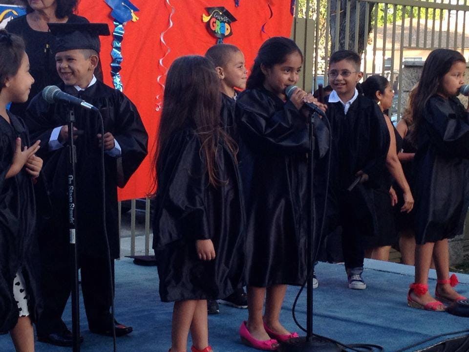 Children in graduation gowns singing into microphones on a stage