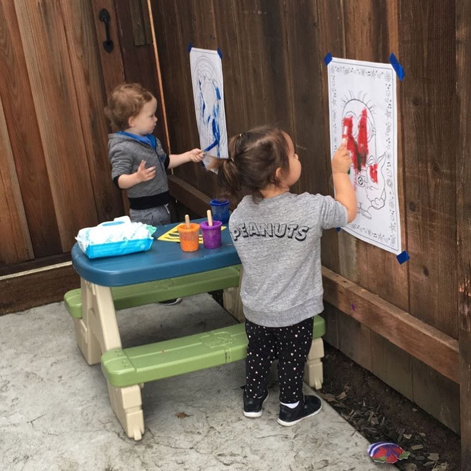 Two children painting on paper attached to a wooden fence