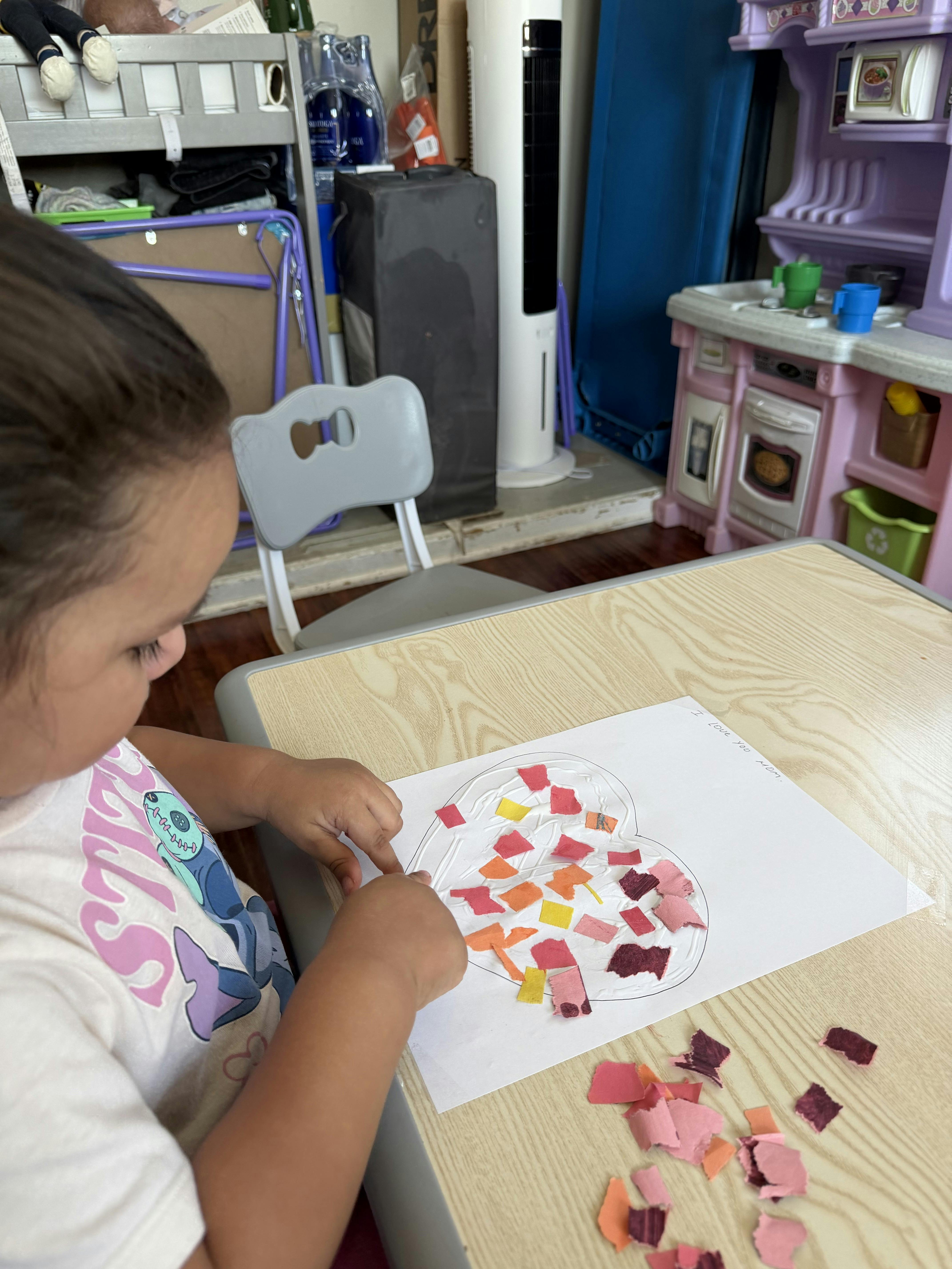 Child making a heart collage with colorful paper scraps and glue on a desk