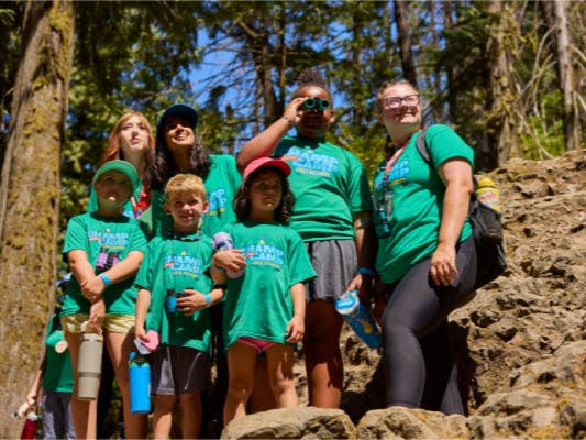 Group of children and adults in green shirts outdoors, one child looking through binoculars