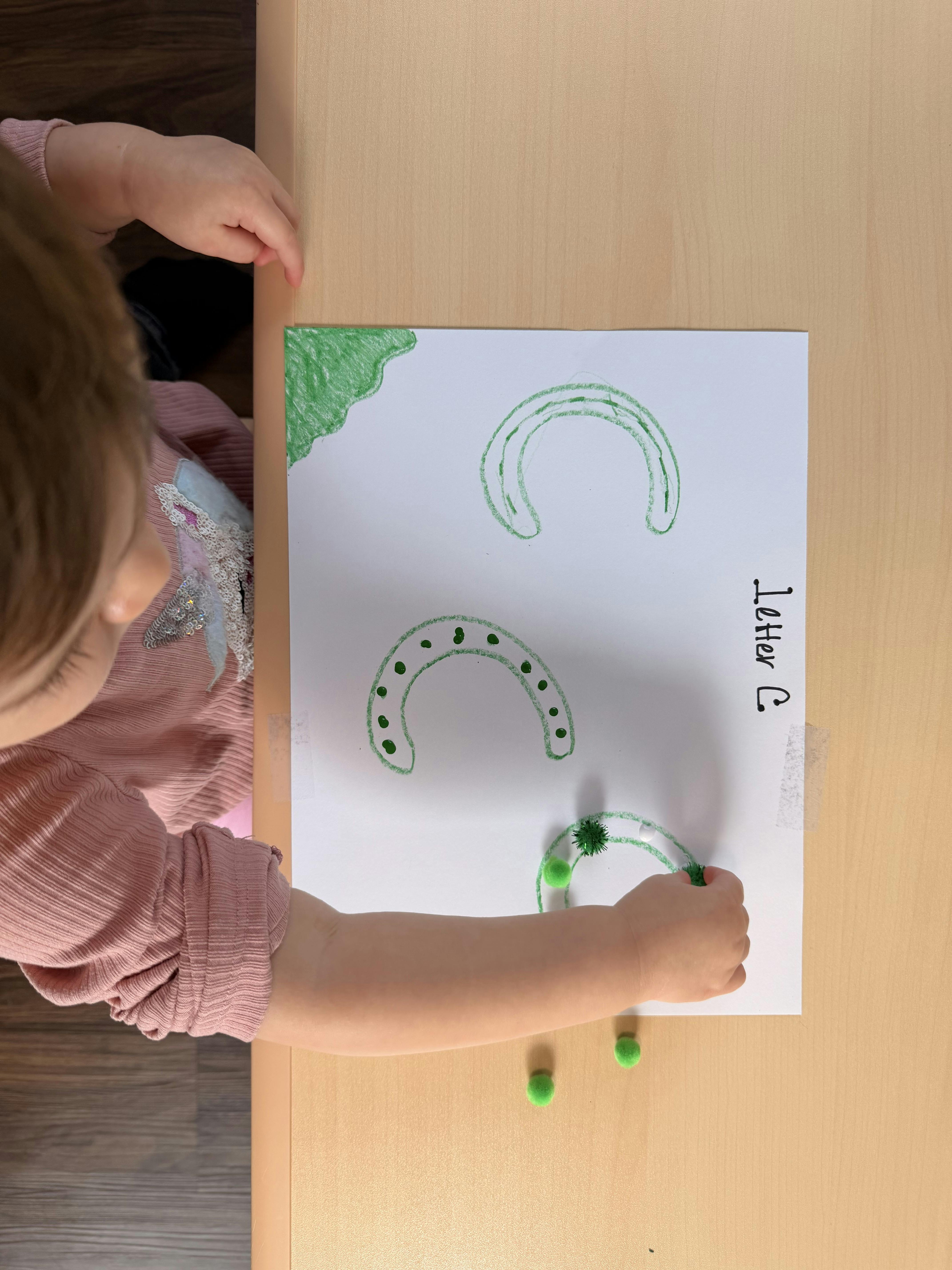 Child placing green pom poms onto a paper with outlined letter C shapes