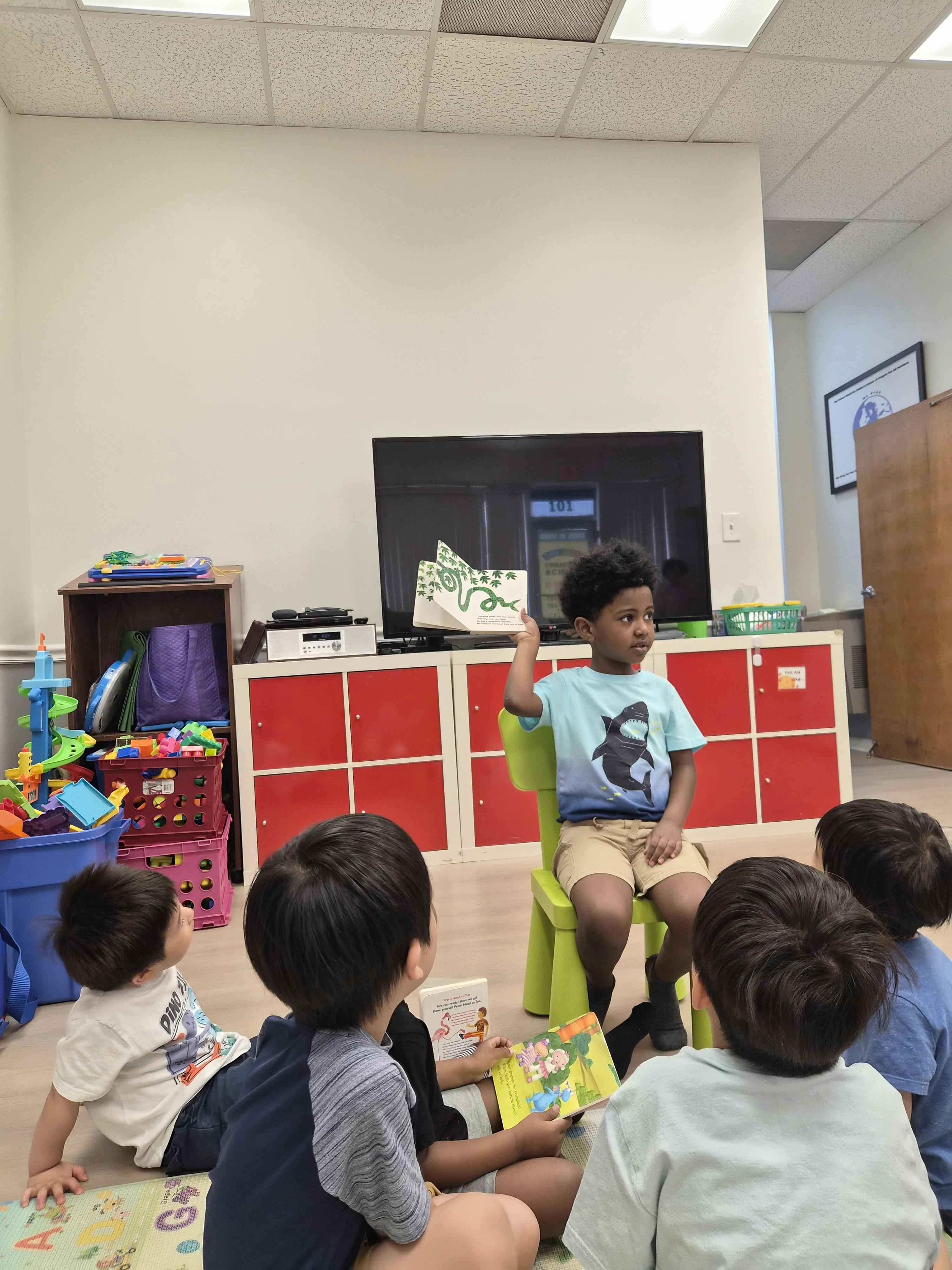 Child sitting on a chair reading a book to a group of children in a classroom