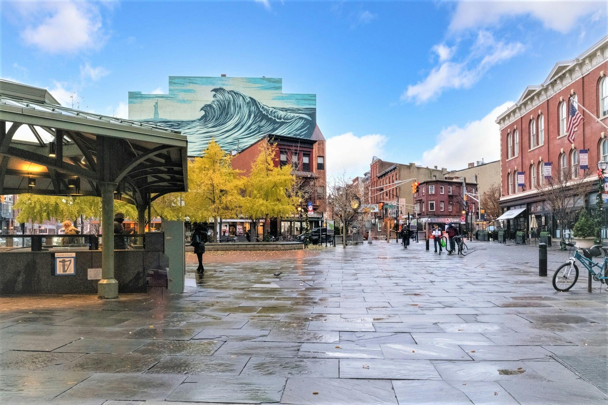People walking on a wet city street with buildings and a wave mural