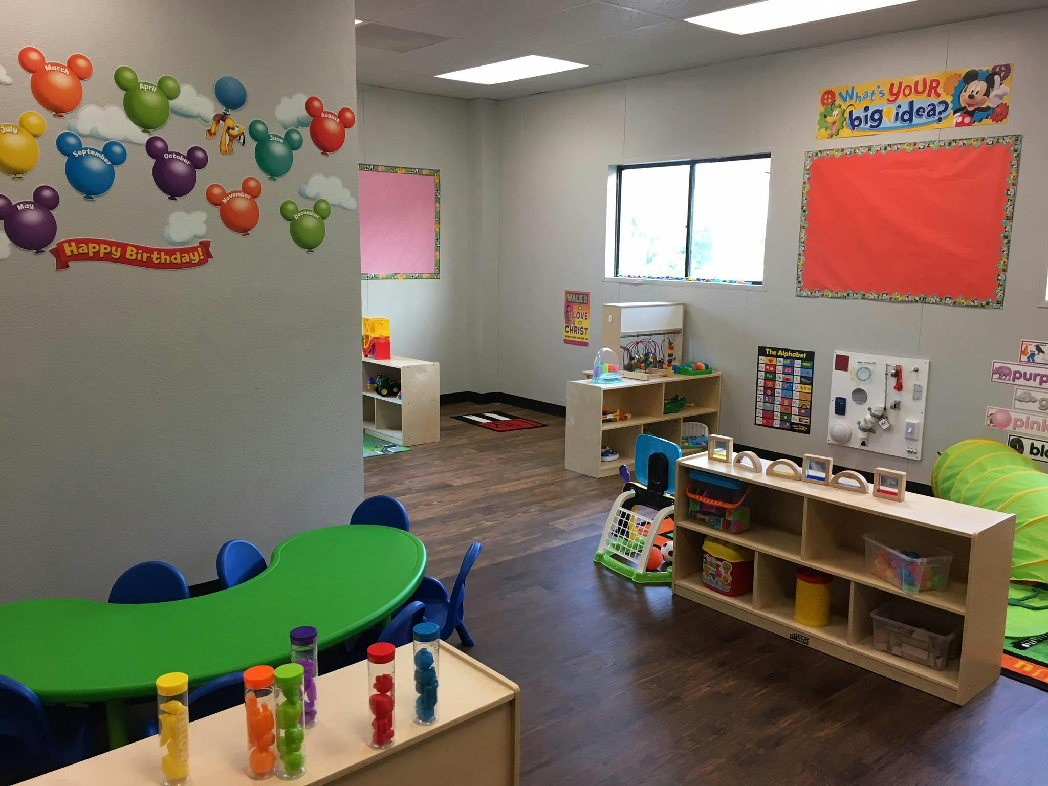 Playroom with small green tables and chairs, shelves with toys, and bulletin boards