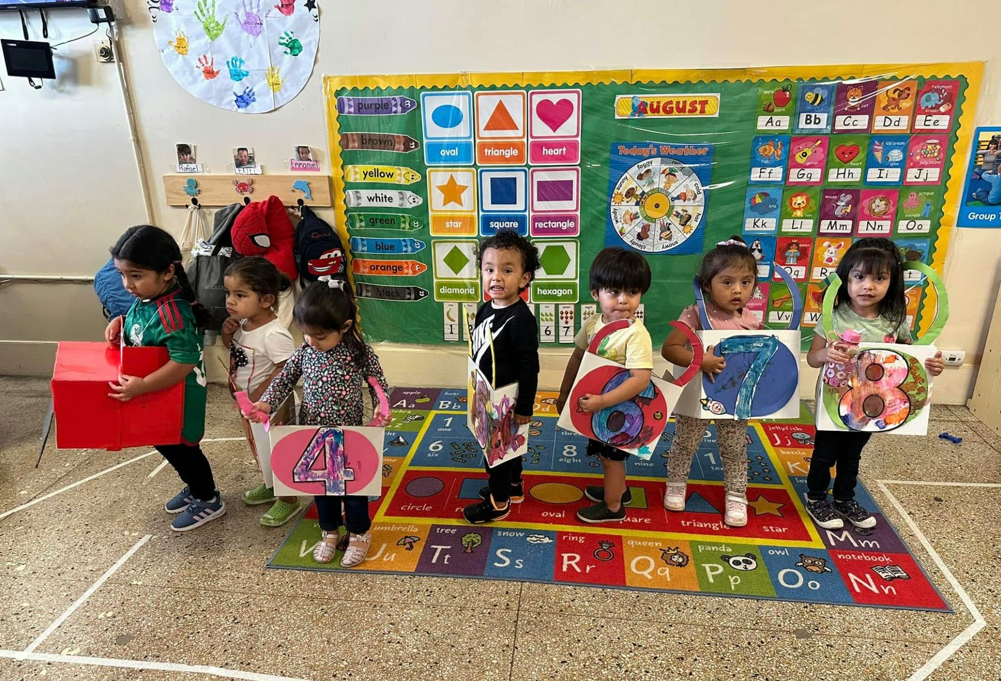 Children in a classroom holding up number cutouts, with a learning chart in the background