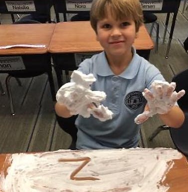 Child with hands covered in white foam, a letter Z drawn in foam on a desk