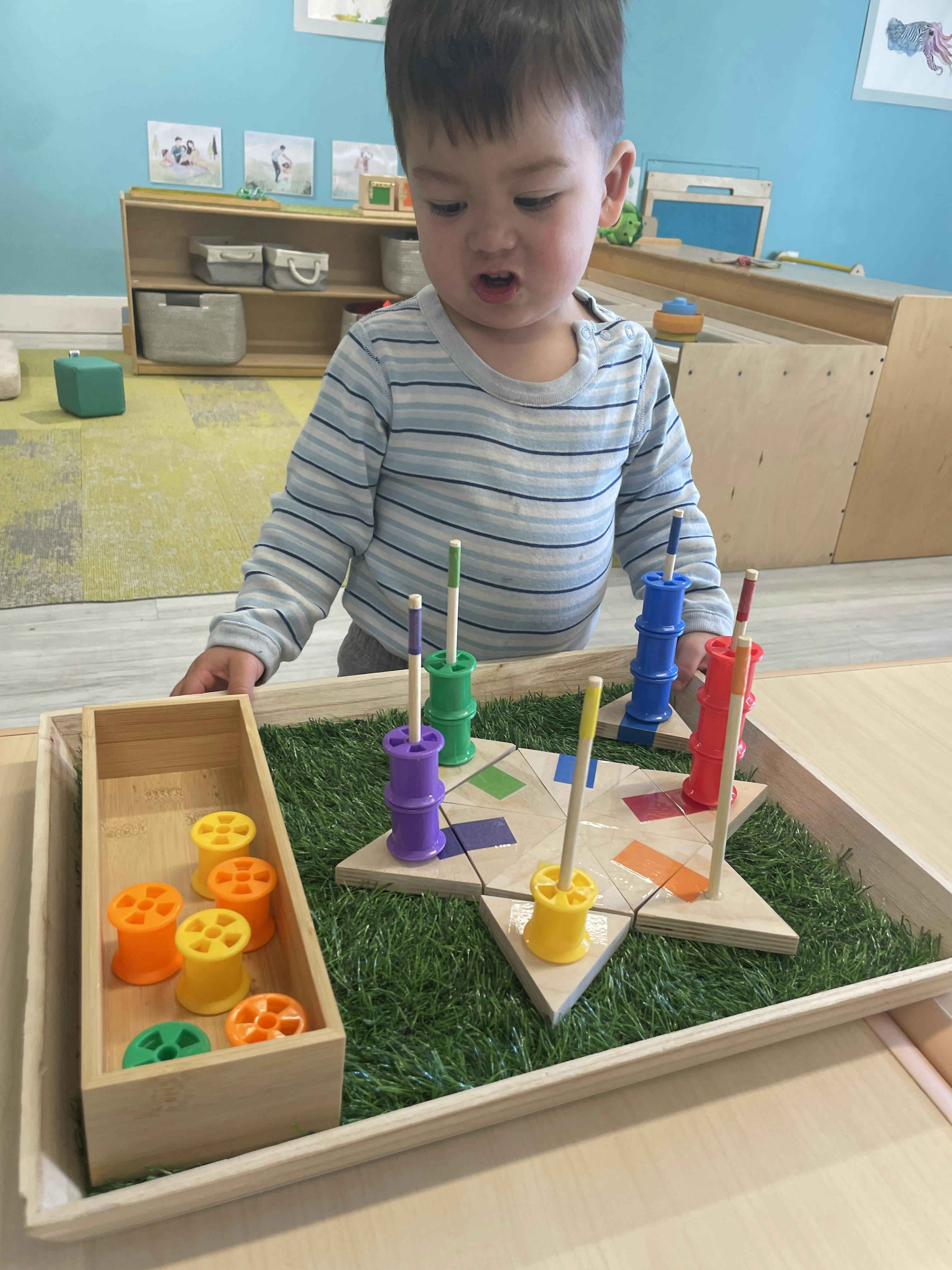 Child playing with a colorful peg and spool toy on a tray with artificial grass