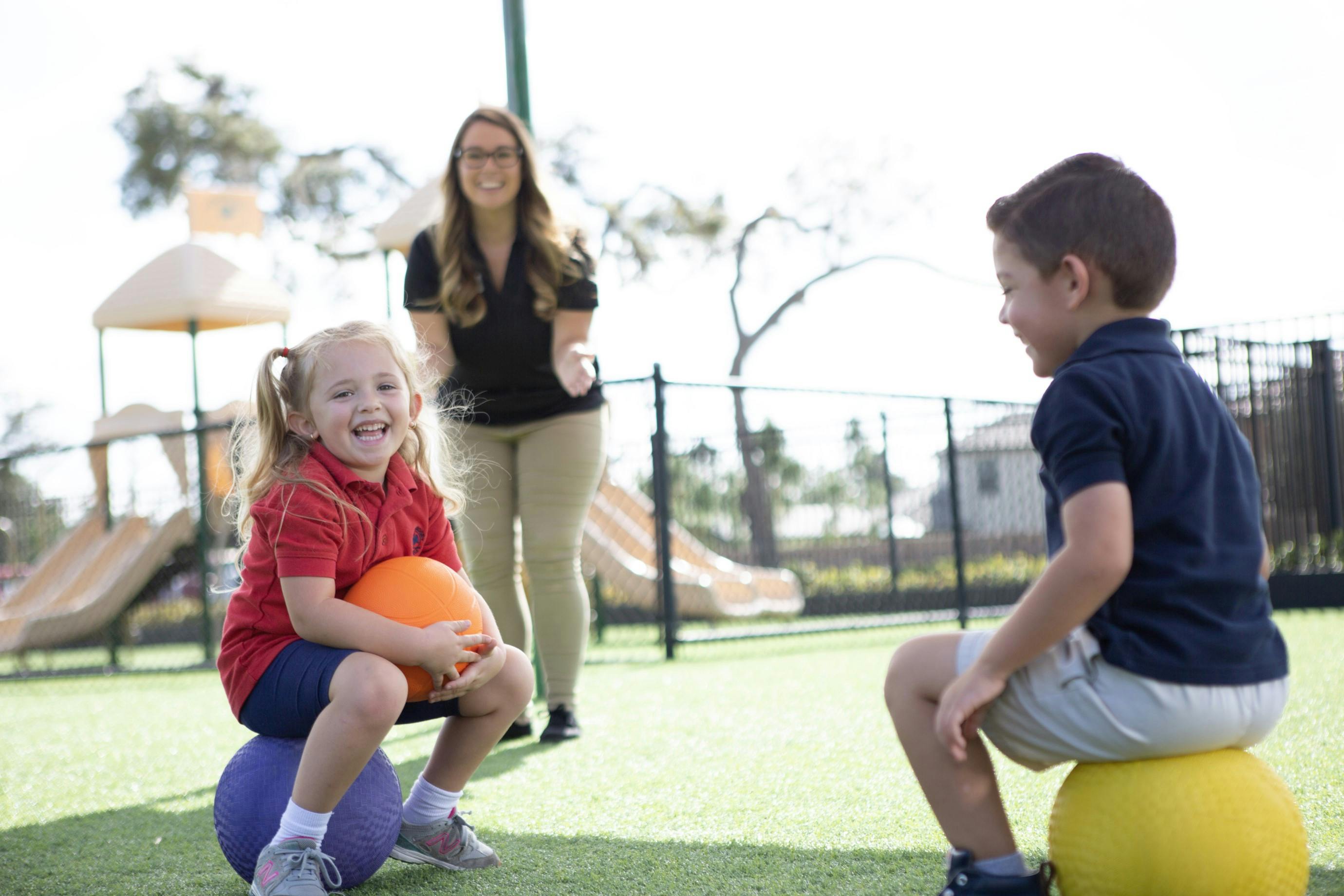 Two children sitting on balls with a person standing behind them on a playground