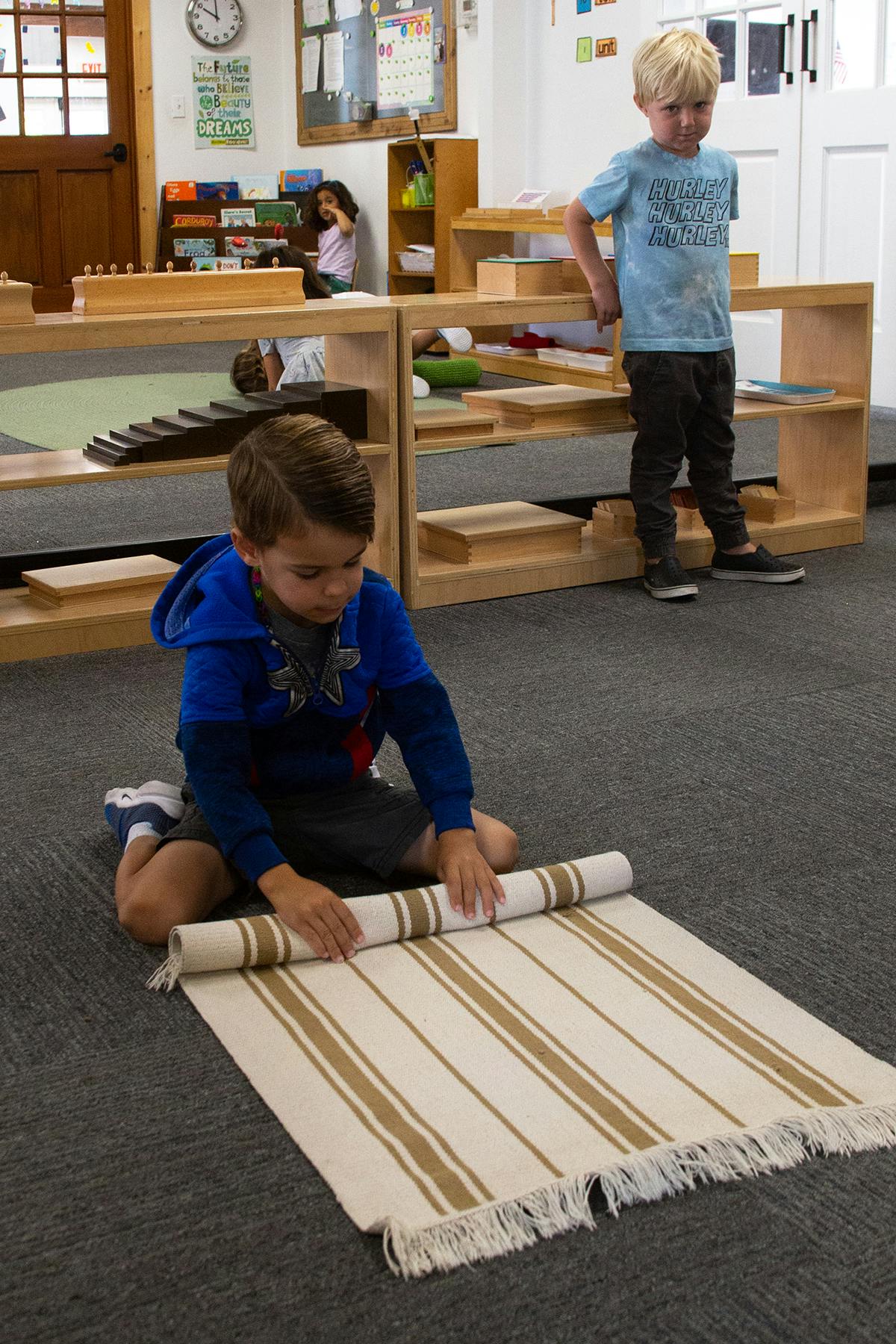 Child kneeling on floor rolling up a striped mat in a classroom