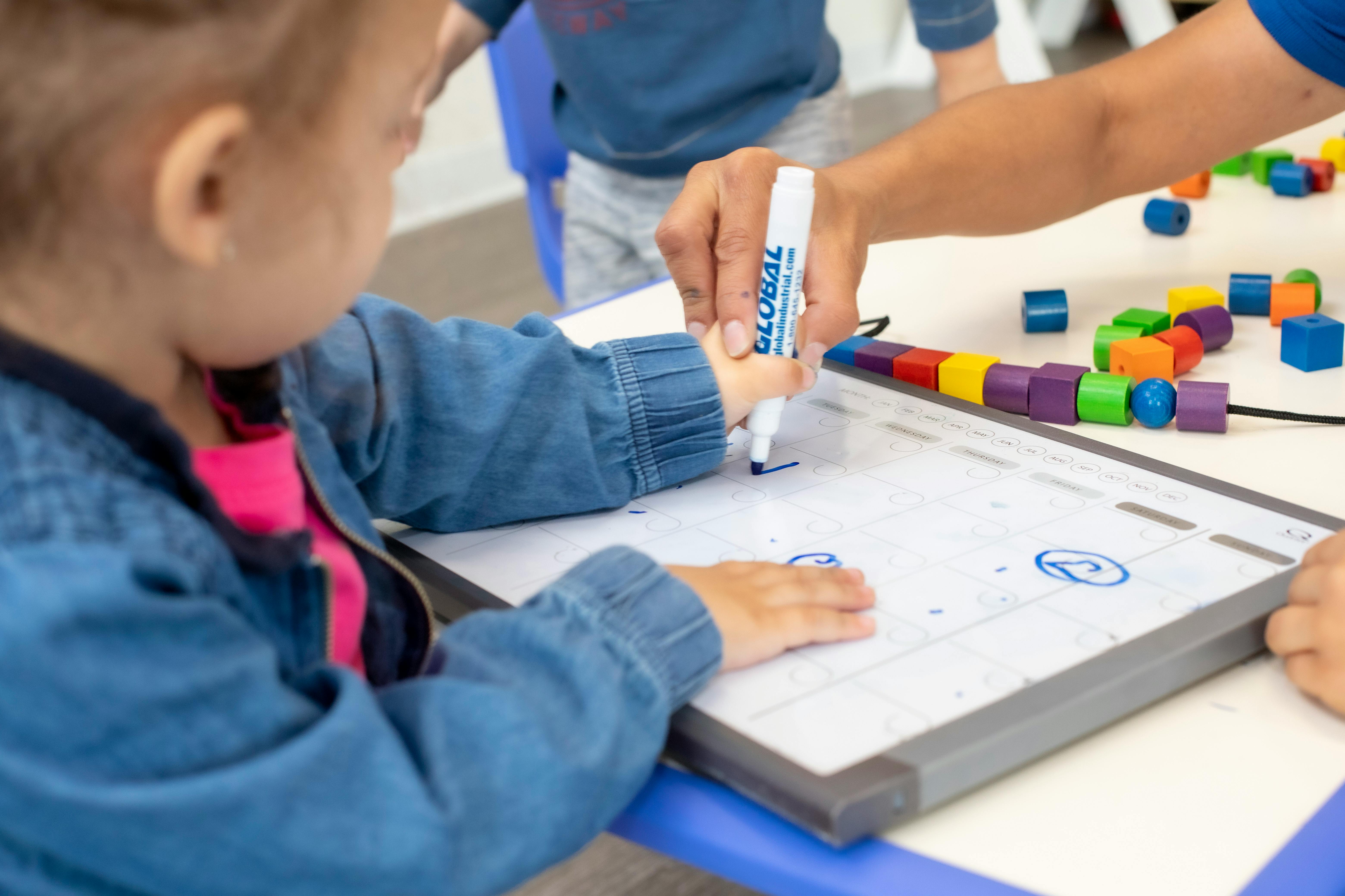 Adult hand helping a child draw on a white board with colorful blocks nearby