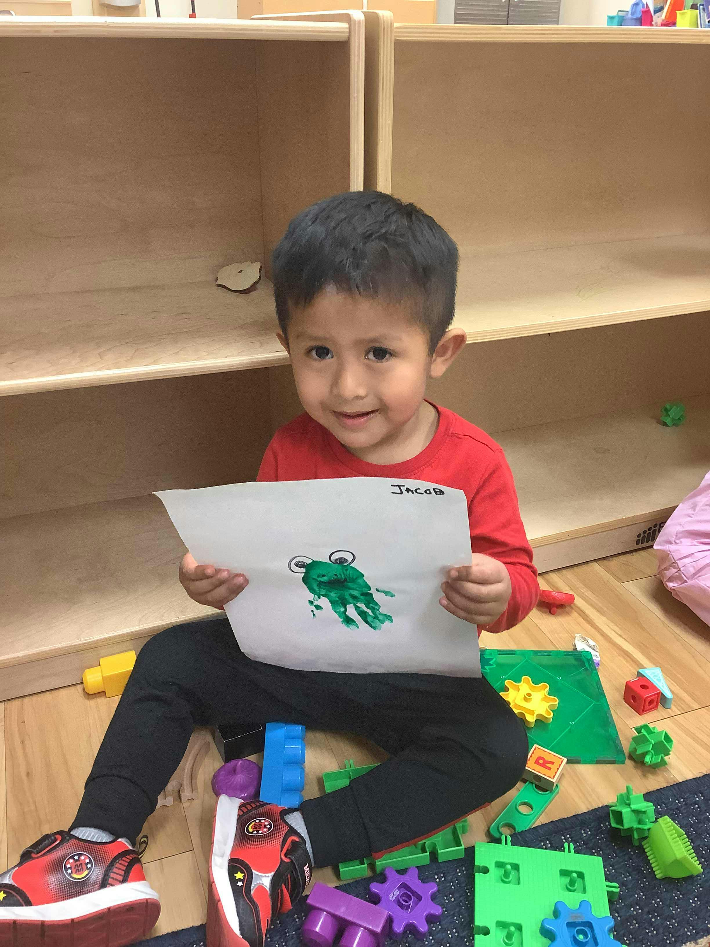 Child sitting on floor holding up a drawing of a frog