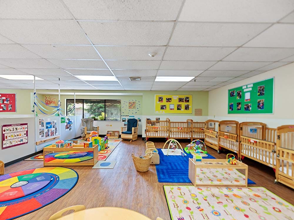 a brightly lit nursery room with multiple cribs, rugs, and play items