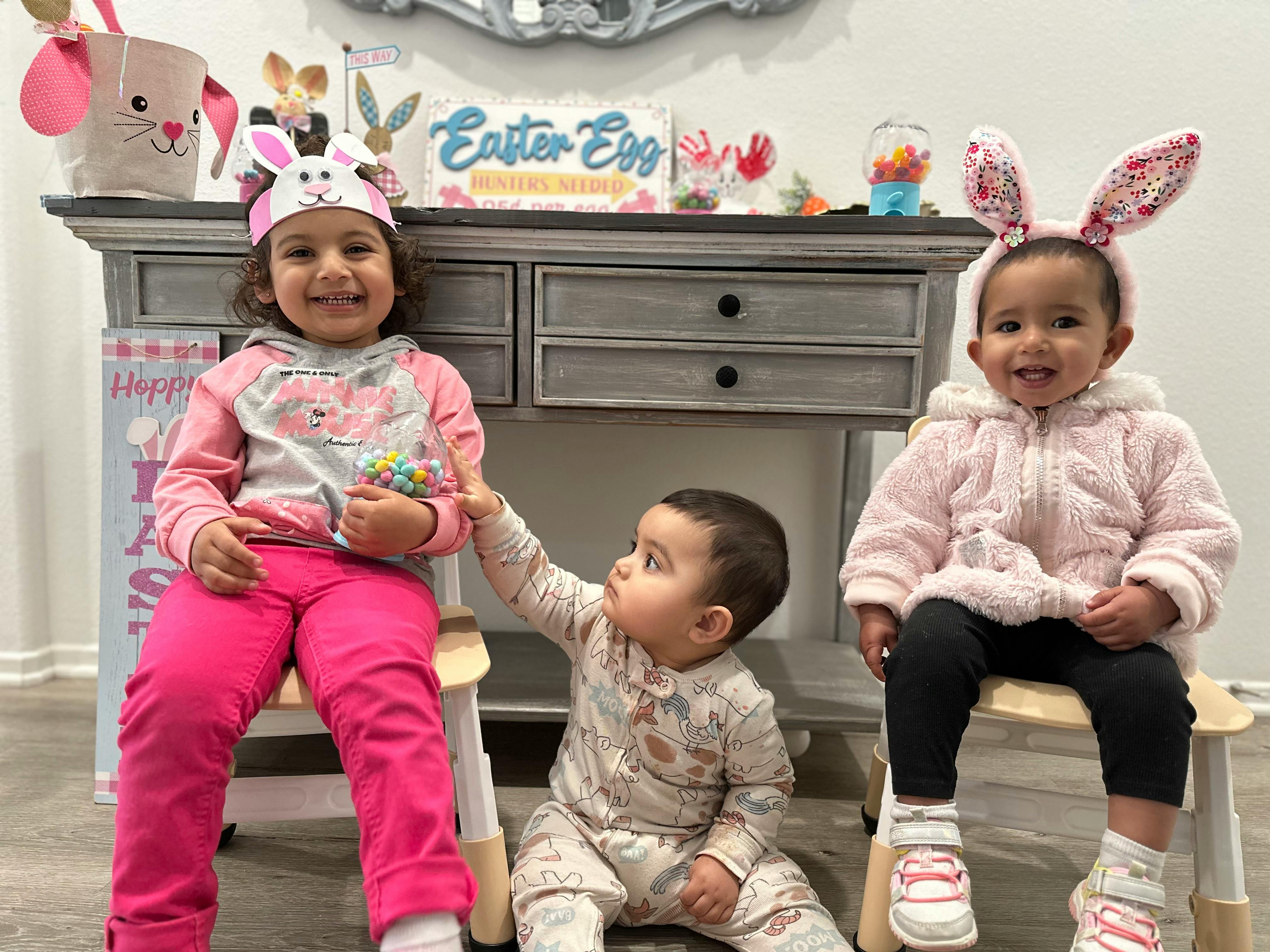Three children, two wearing bunny ears, one sitting on a chair, one on the floor