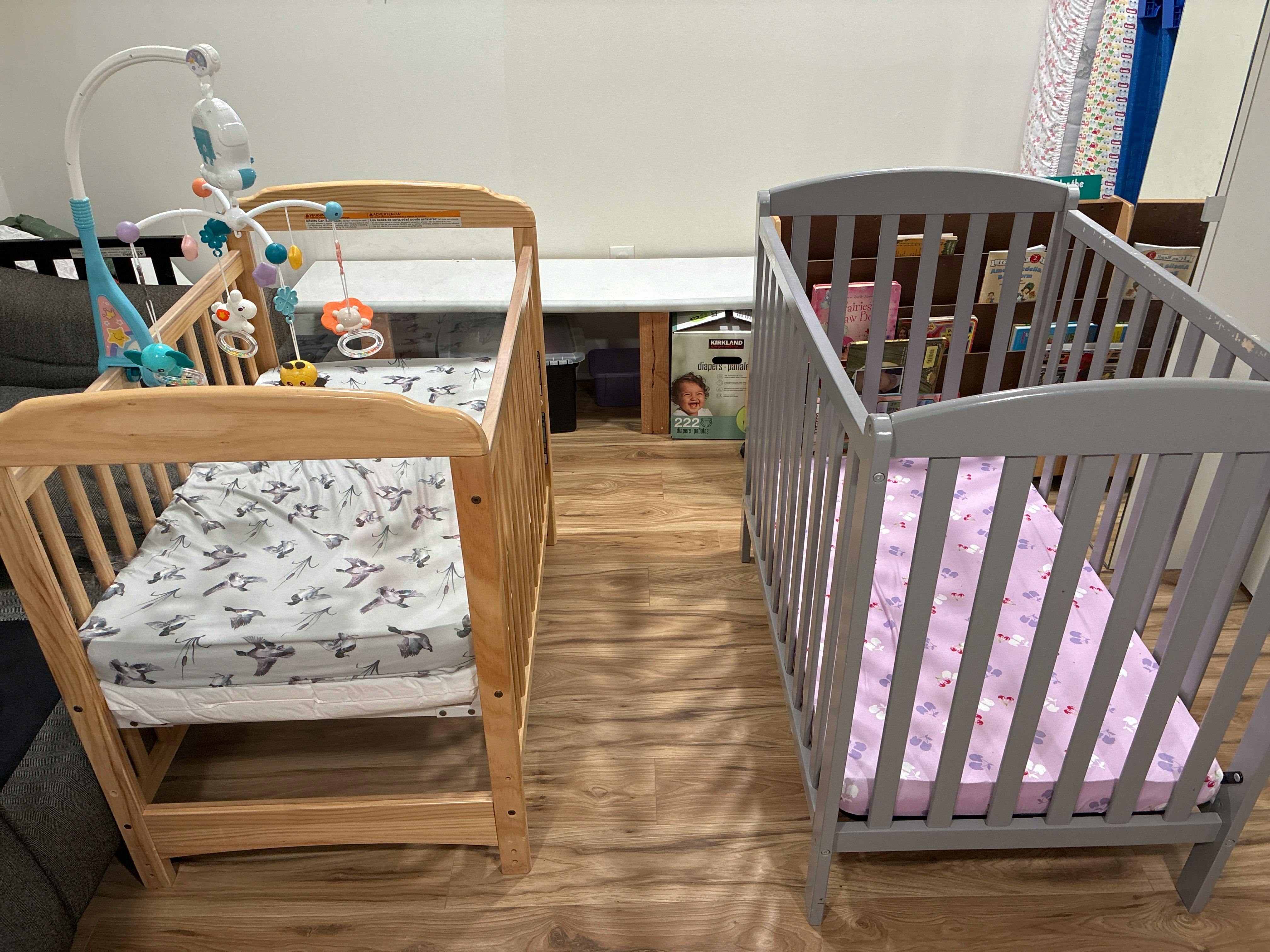 two empty cribs side by side, one wooden with a mobile, one gray