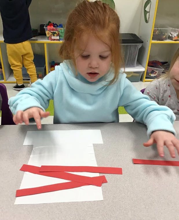 Child working on a craft with red paper strips on a white paper shape