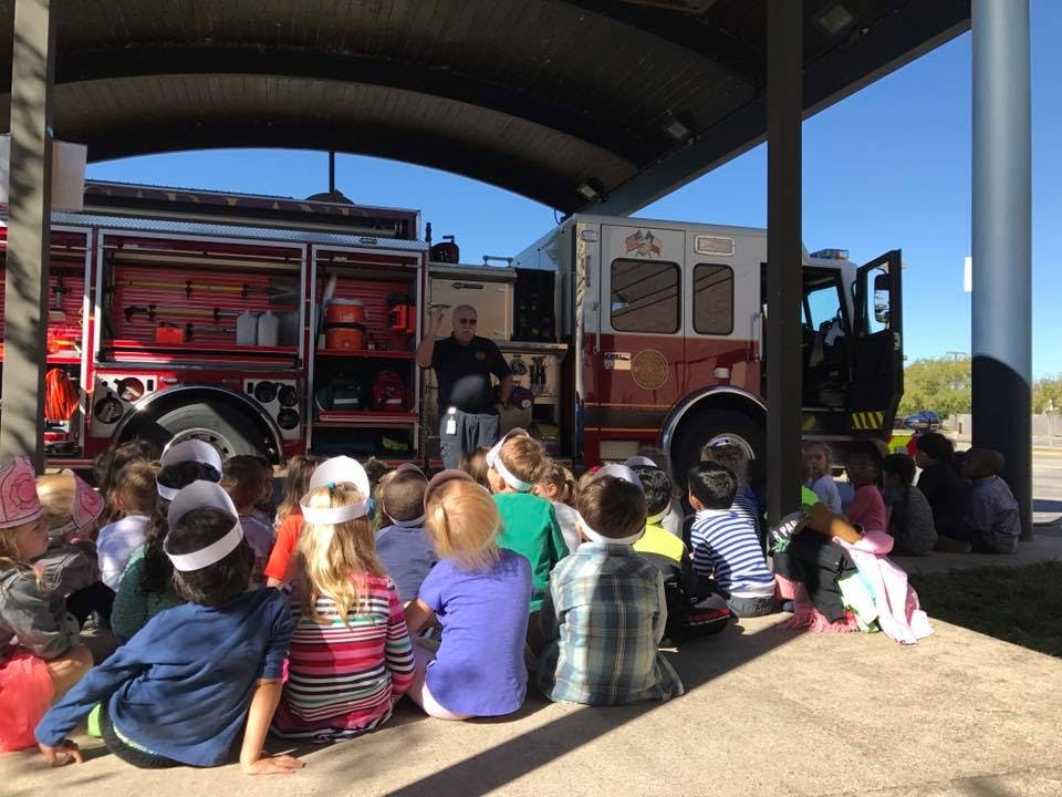 Children sitting on the ground in front of a fire truck with an adult