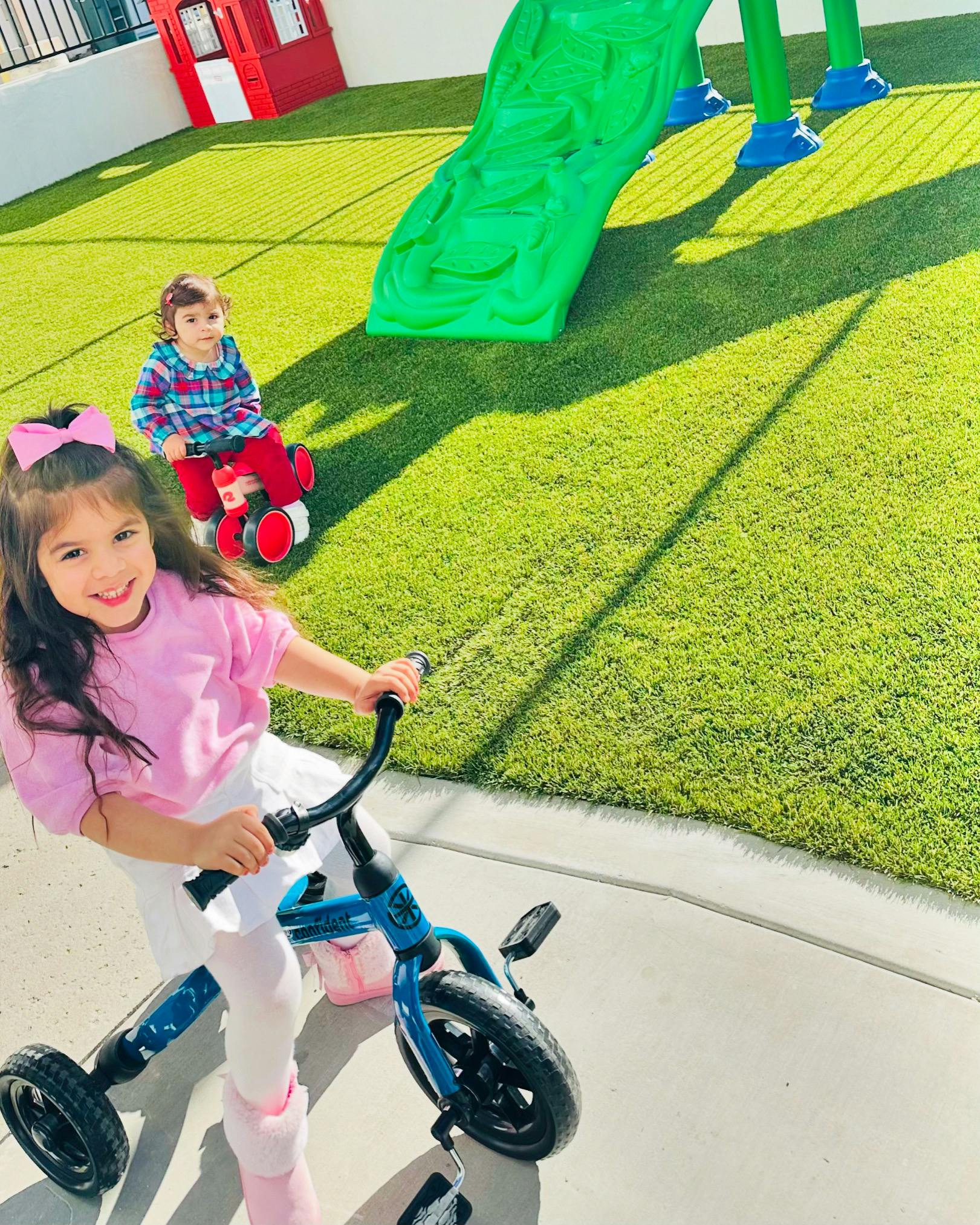 Child on a tricycle on a sidewalk, another child on a ride-on toy in background