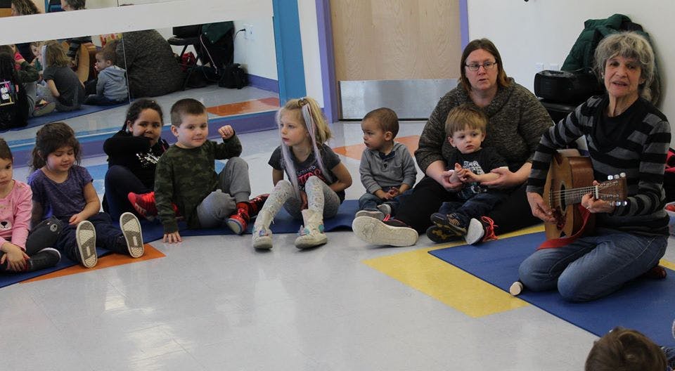 Adults and children sitting on the floor, one adult playing a guitar
