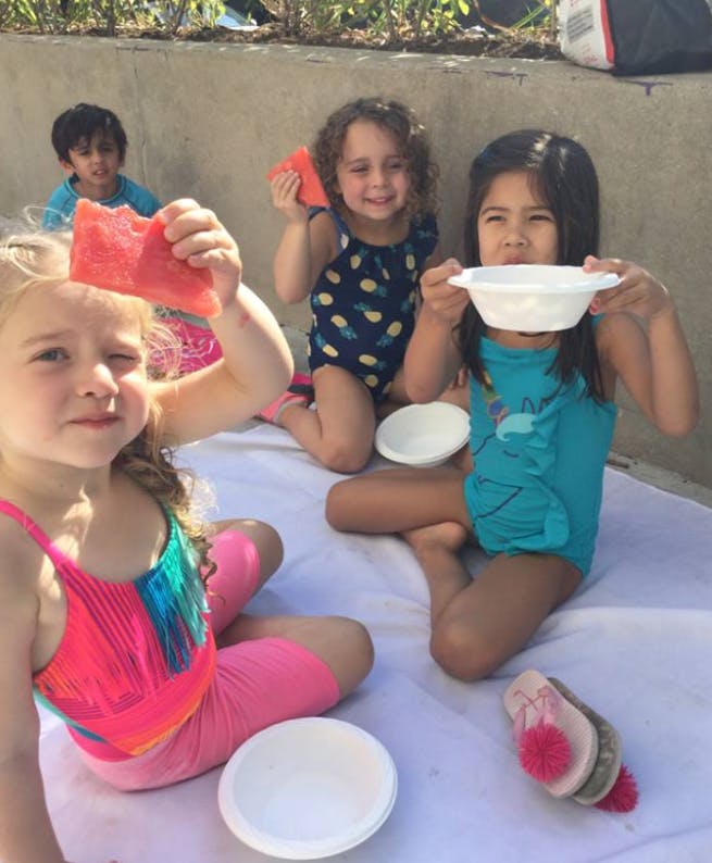 Three children sitting on a blanket, two holding watermelon slices, one holding a bowl