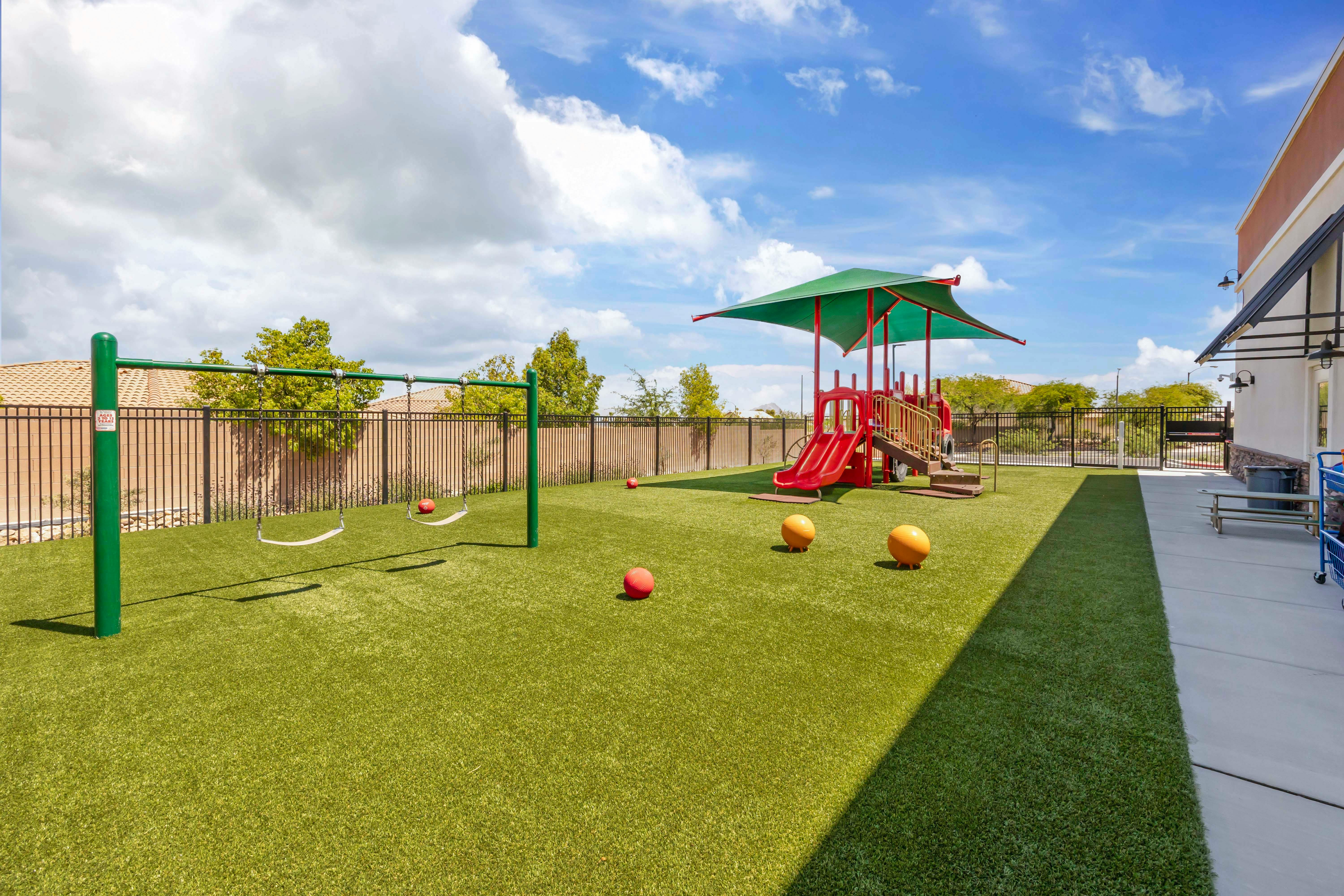 Playground with swings, a red and green play structure, and red balls on artificial turf