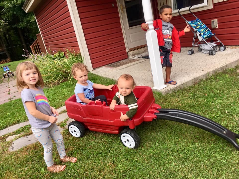 Three children playing with a red wagon outside a red building