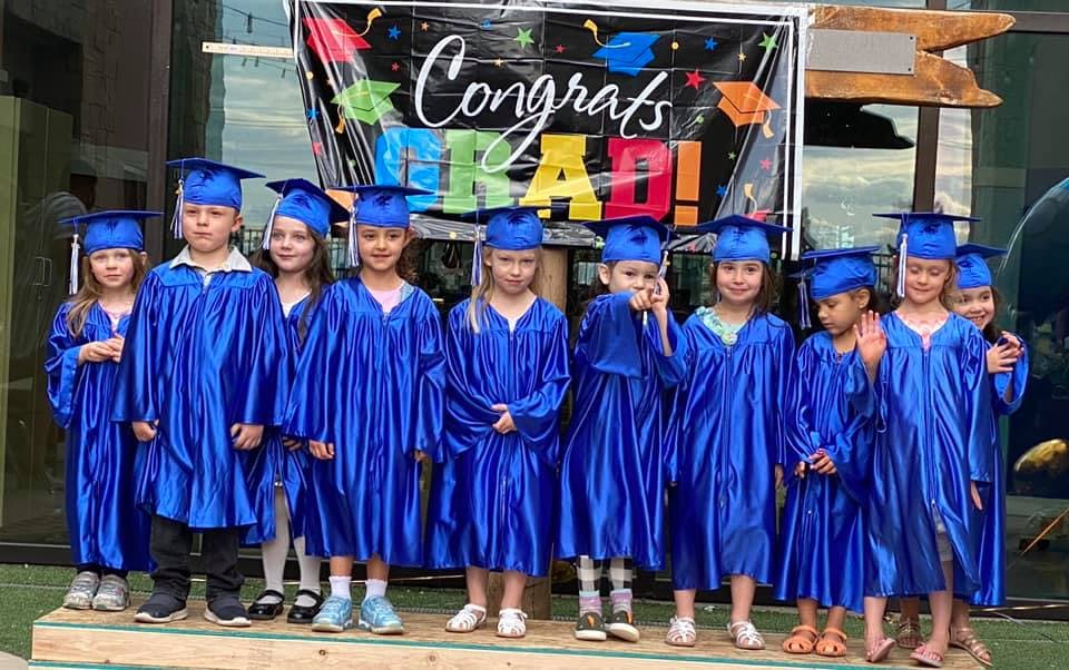 Group of children in blue graduation caps and gowns standing in front of a banner