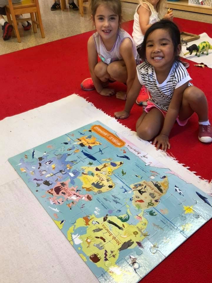 Two children kneeling on a red rug next to a world map puzzle