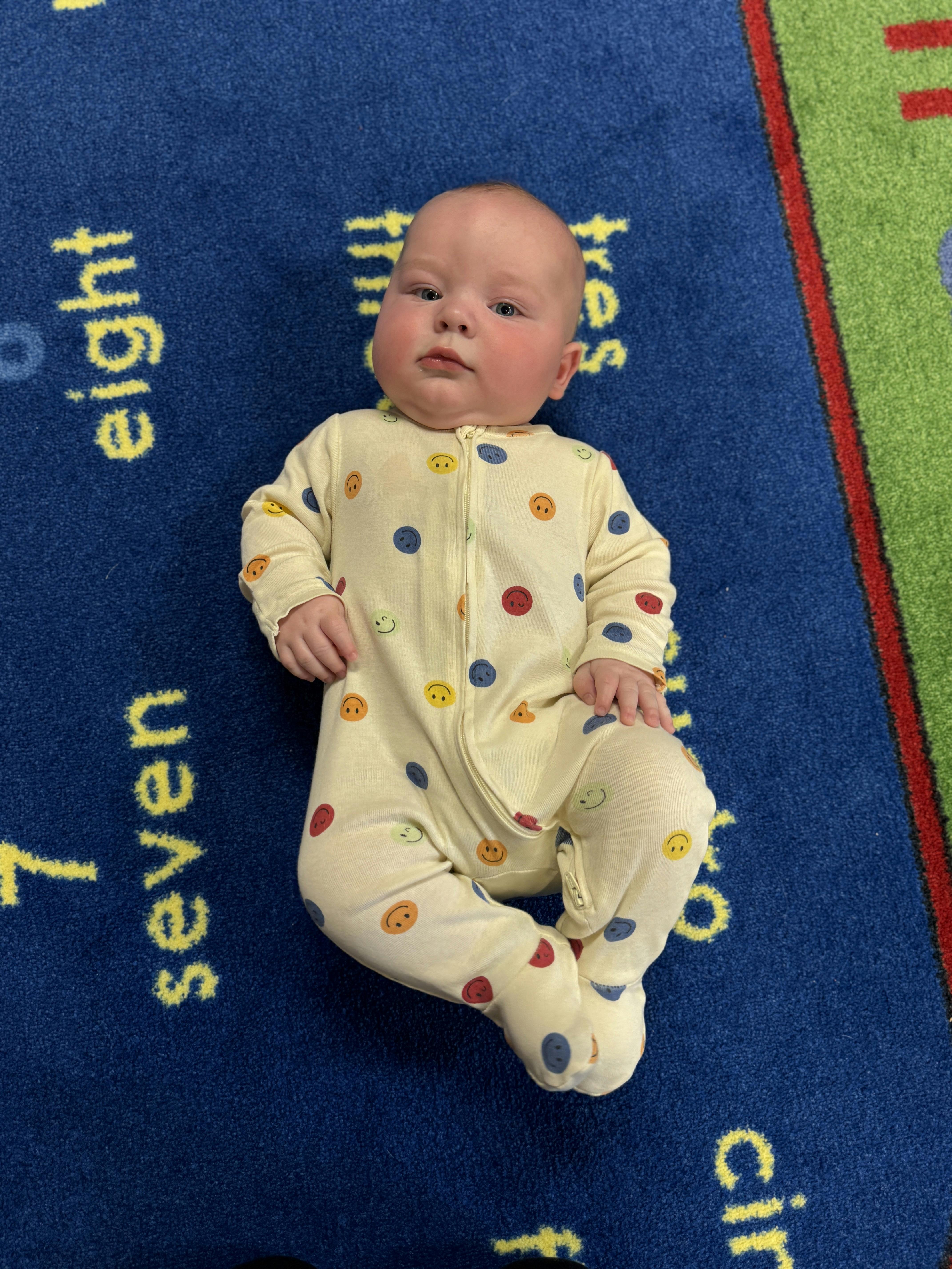 Child in a smiley face onesie lying on a blue rug with numbers