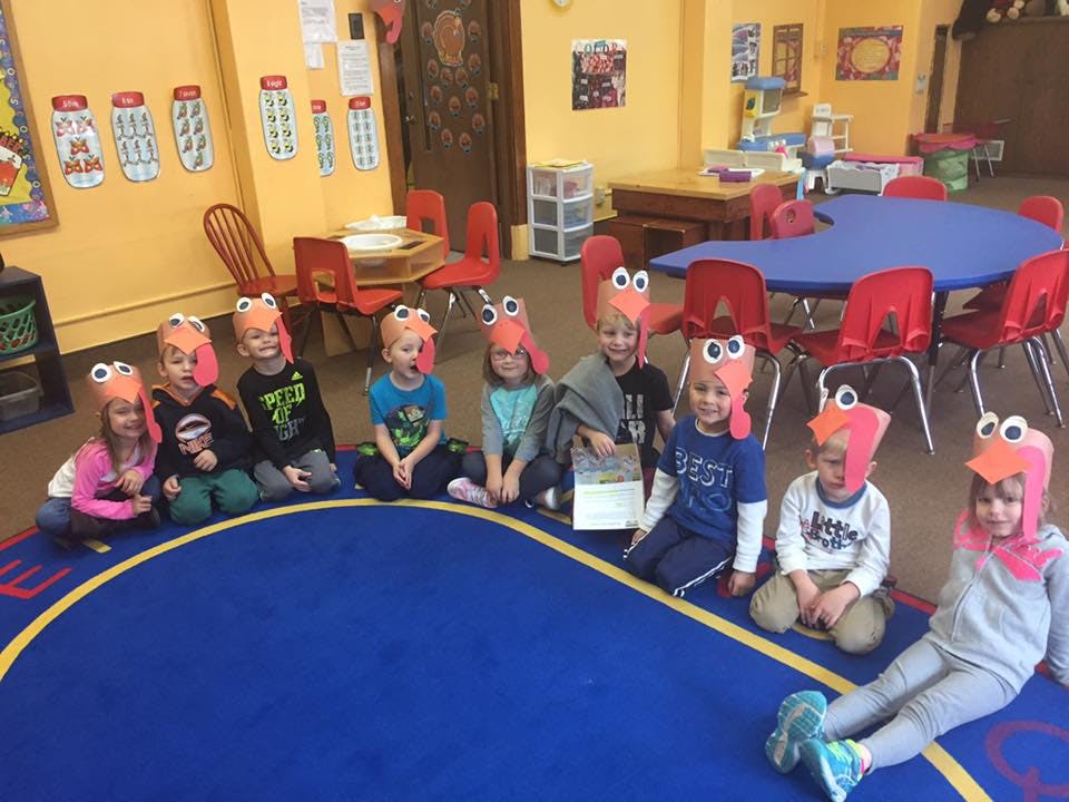 Children wearing bird hats sitting on a blue rug in a classroom