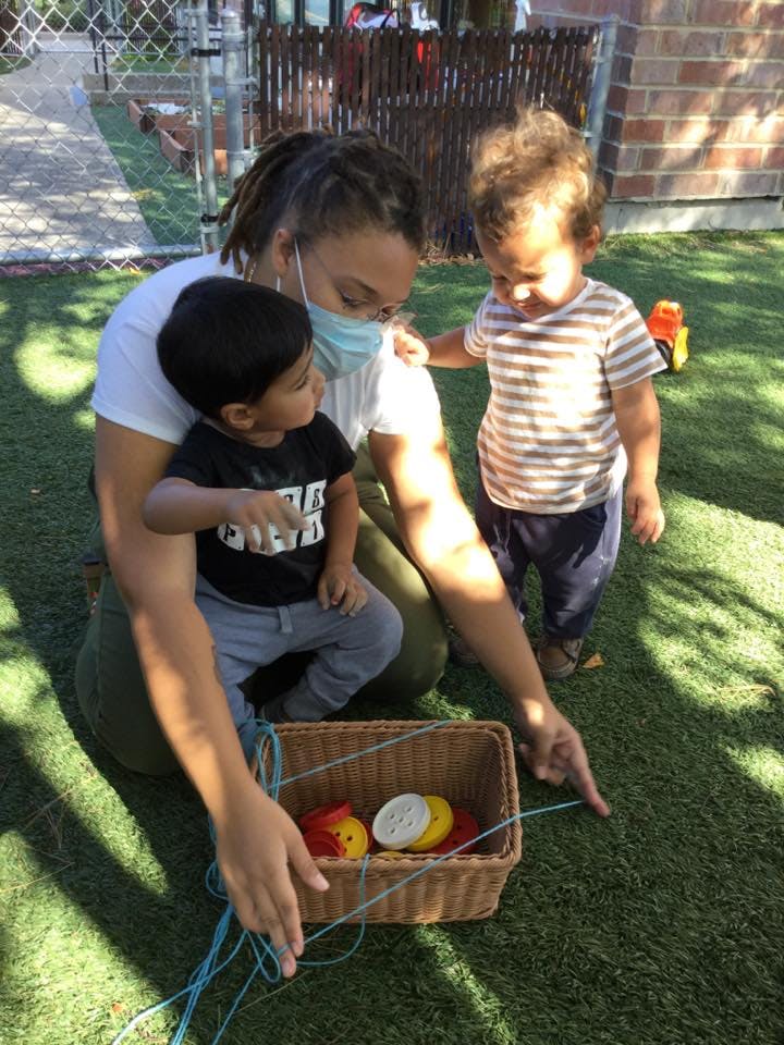 Person in mask with two children on grass, pointing at a basket