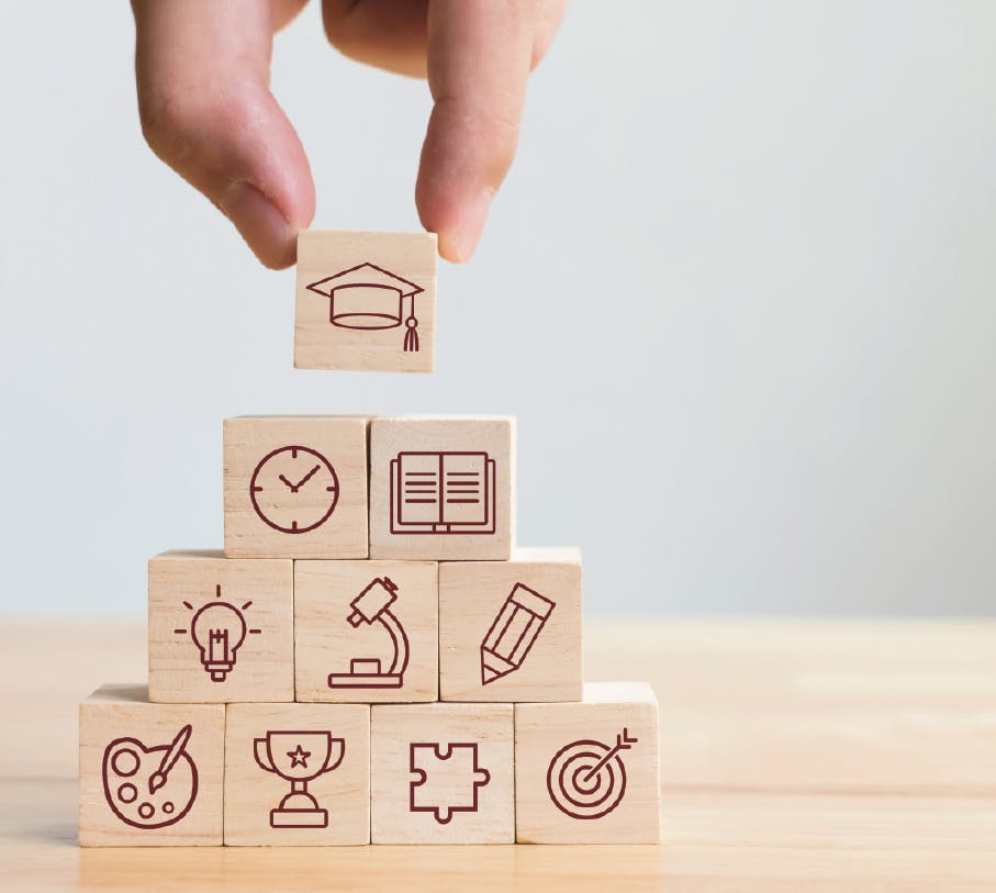 Hand placing a wooden block with a graduation cap on top of a pyramid of blocks