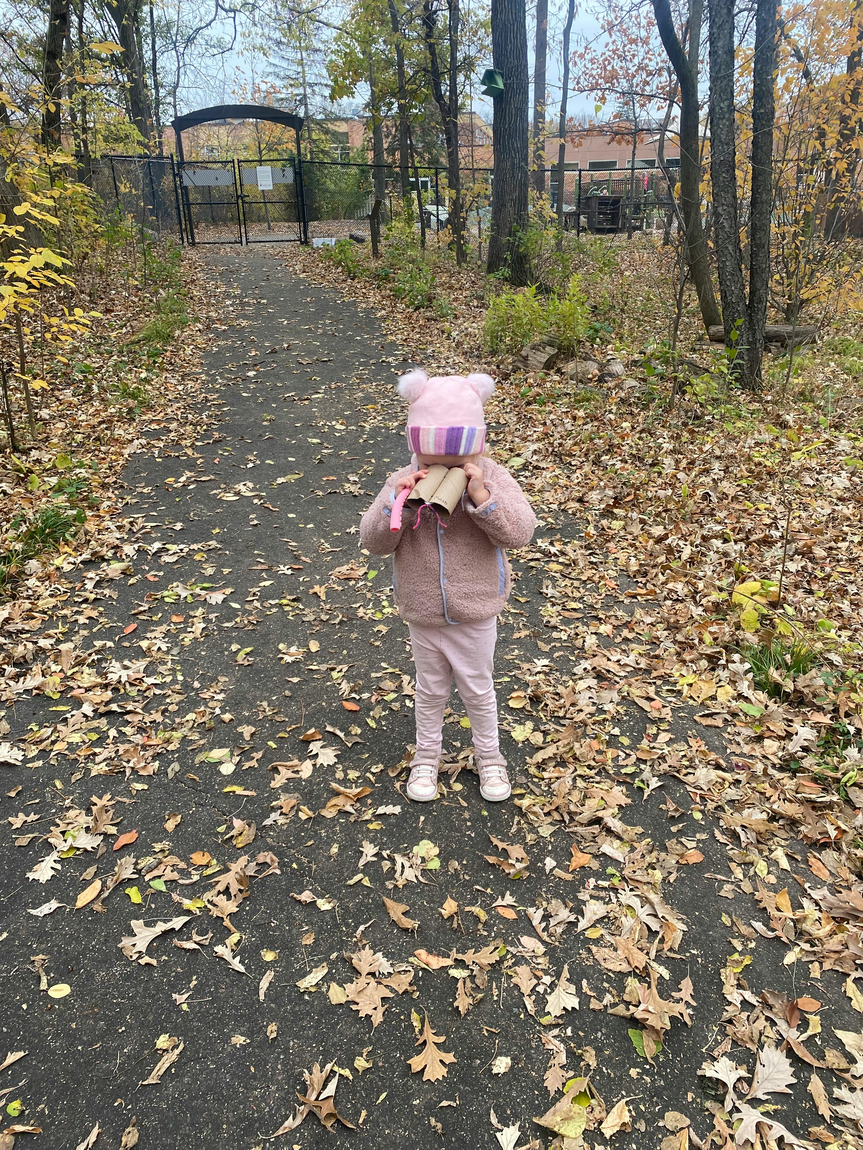 Child in a hat and coat holding paper towel rolls on a leaf-covered path