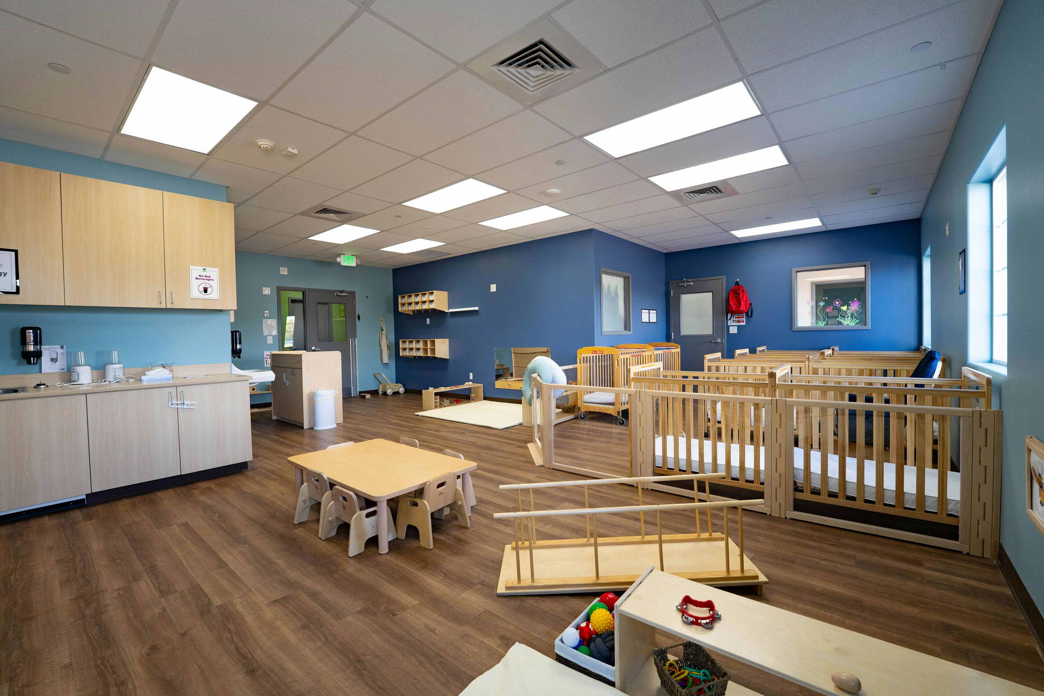 A childcare classroom with cribs, a small table with chairs, and cabinets