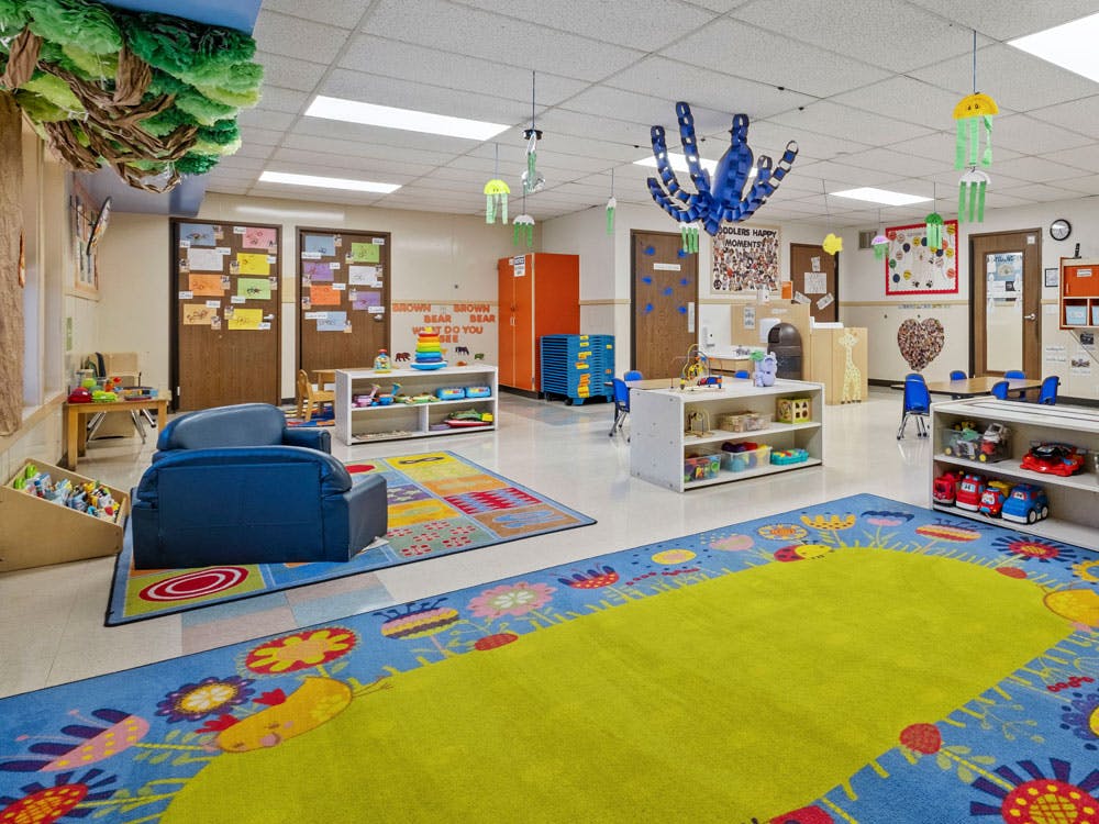 Colorful daycare room with rugs, small tables, chairs, and shelves of toys