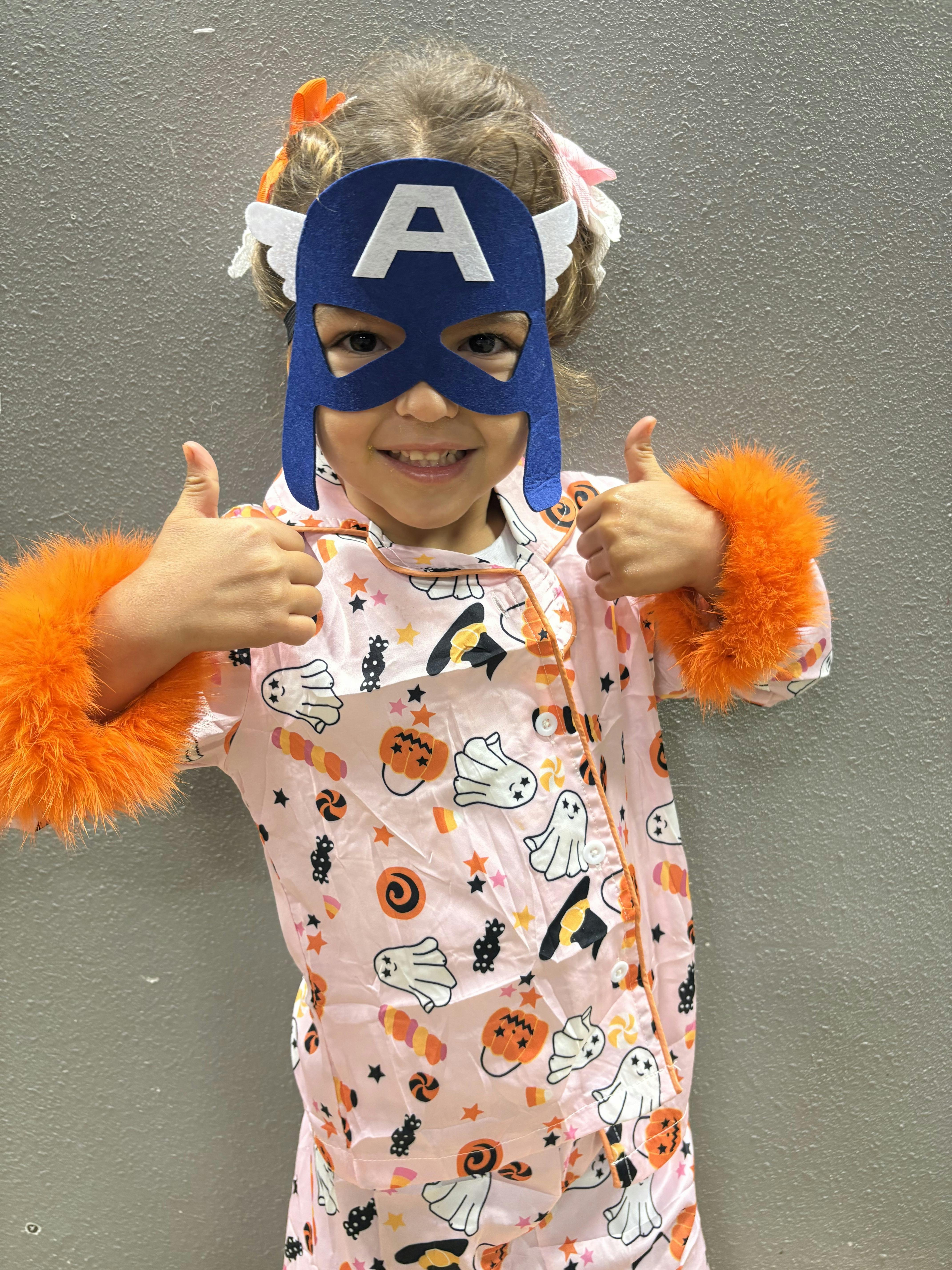 Child in a blue mask with an A and Halloween pajamas giving two thumbs up