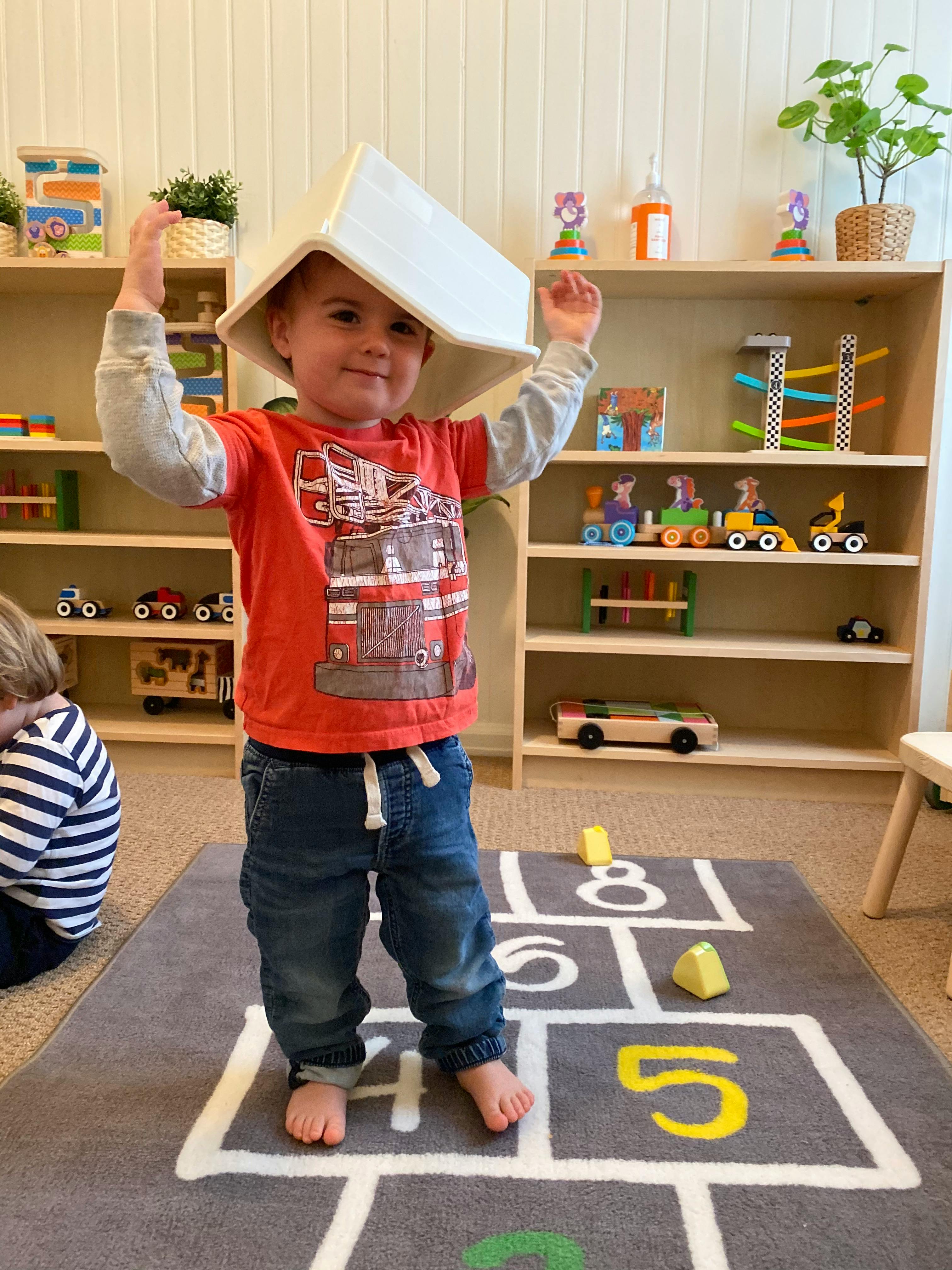 Child with a white box on their head, standing on a hopscotch rug