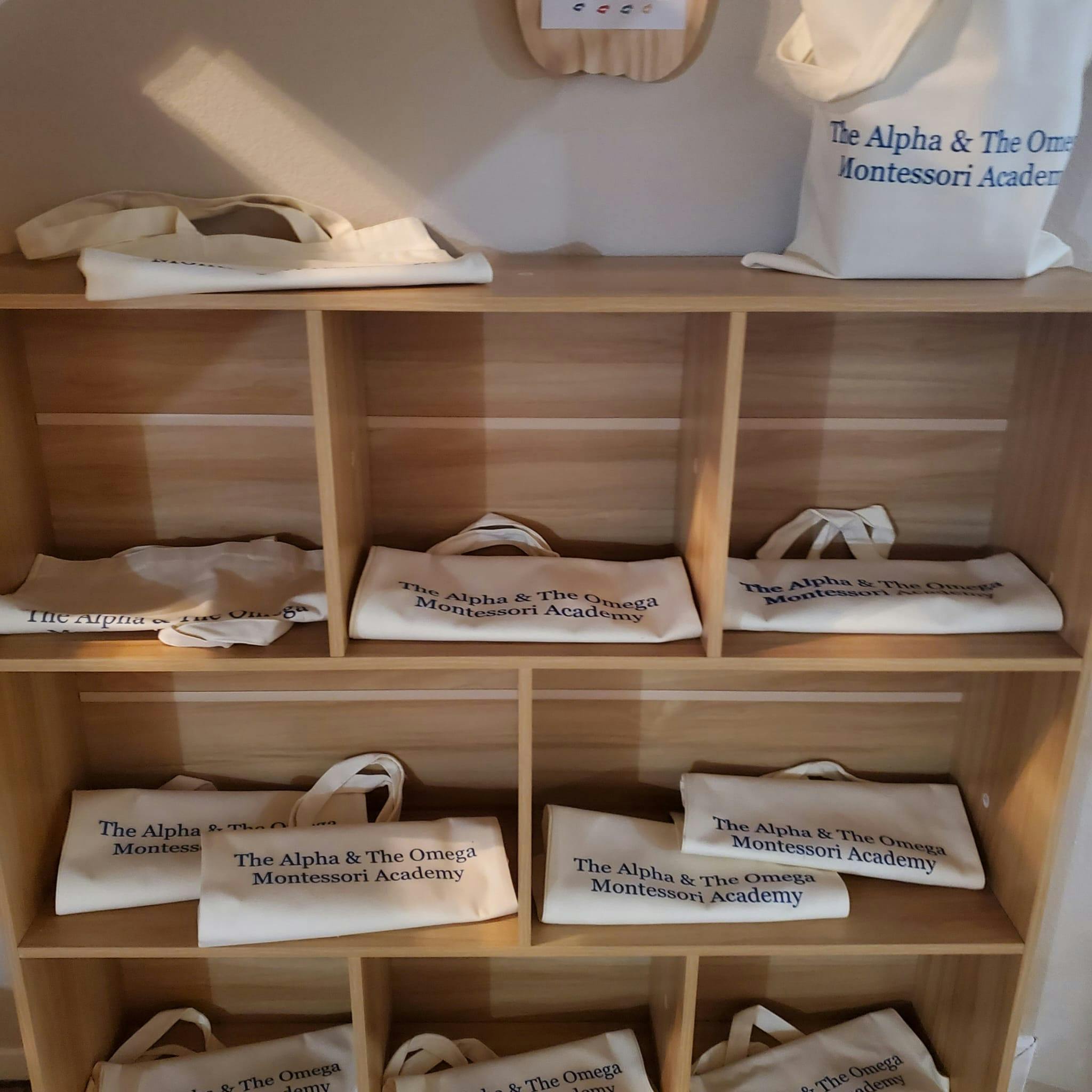 White tote bags with "The Alpha & The Omega Montessori Academy" text on shelves