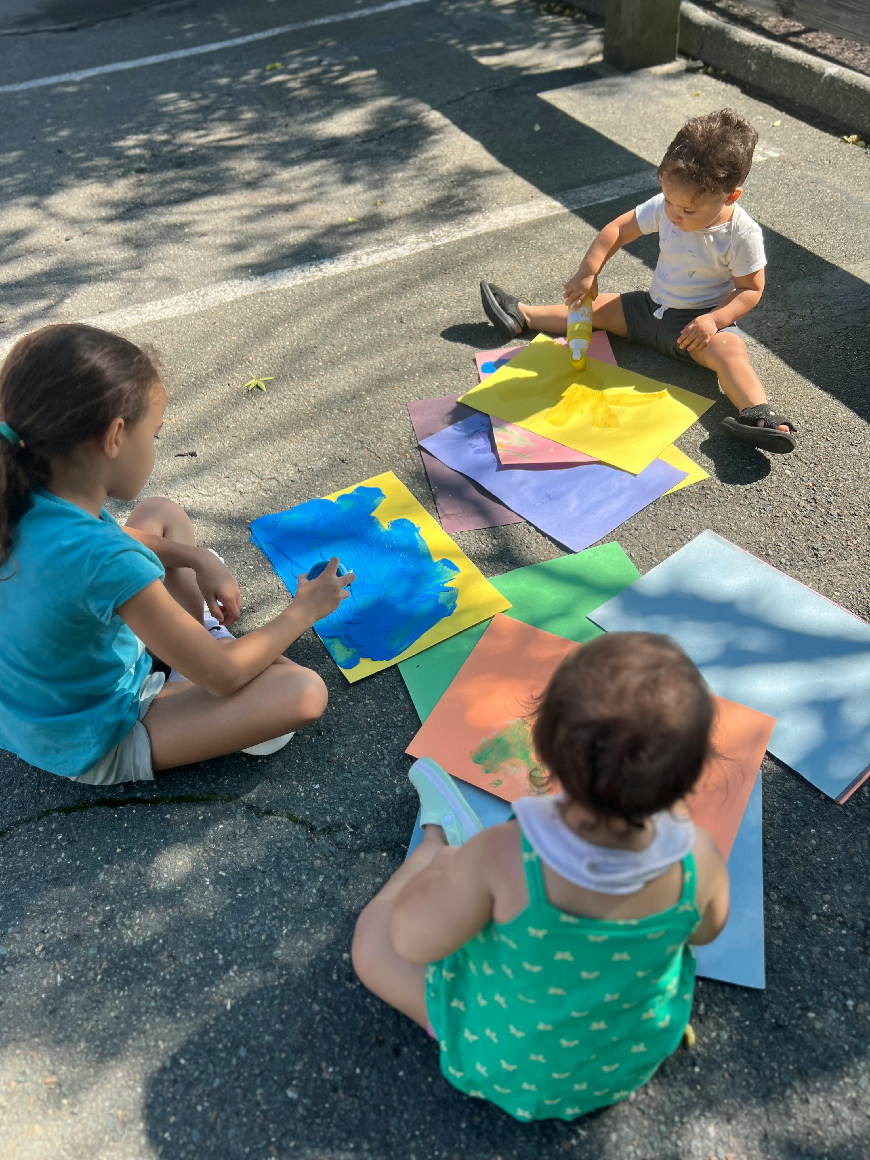 Three children sitting on the ground painting on colorful paper