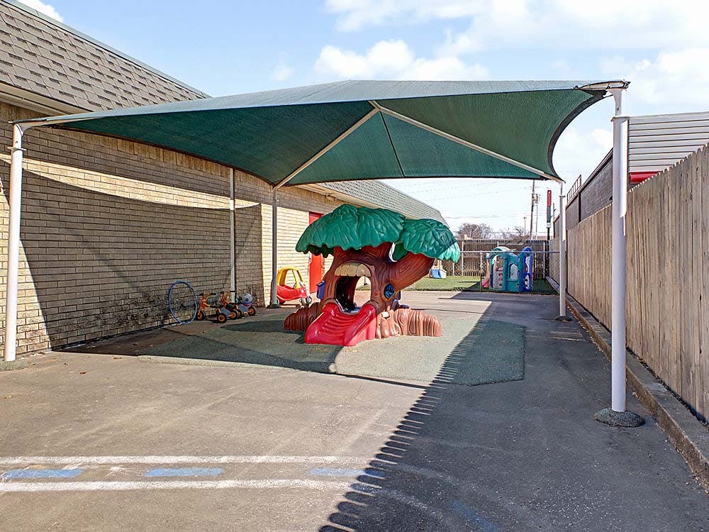 Outdoor play area with a green shade canopy over a tree-shaped slide and trikes