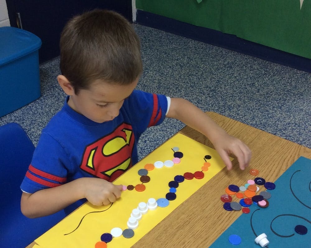 Child placing colorful bottle caps on a yellow paper in a classroom