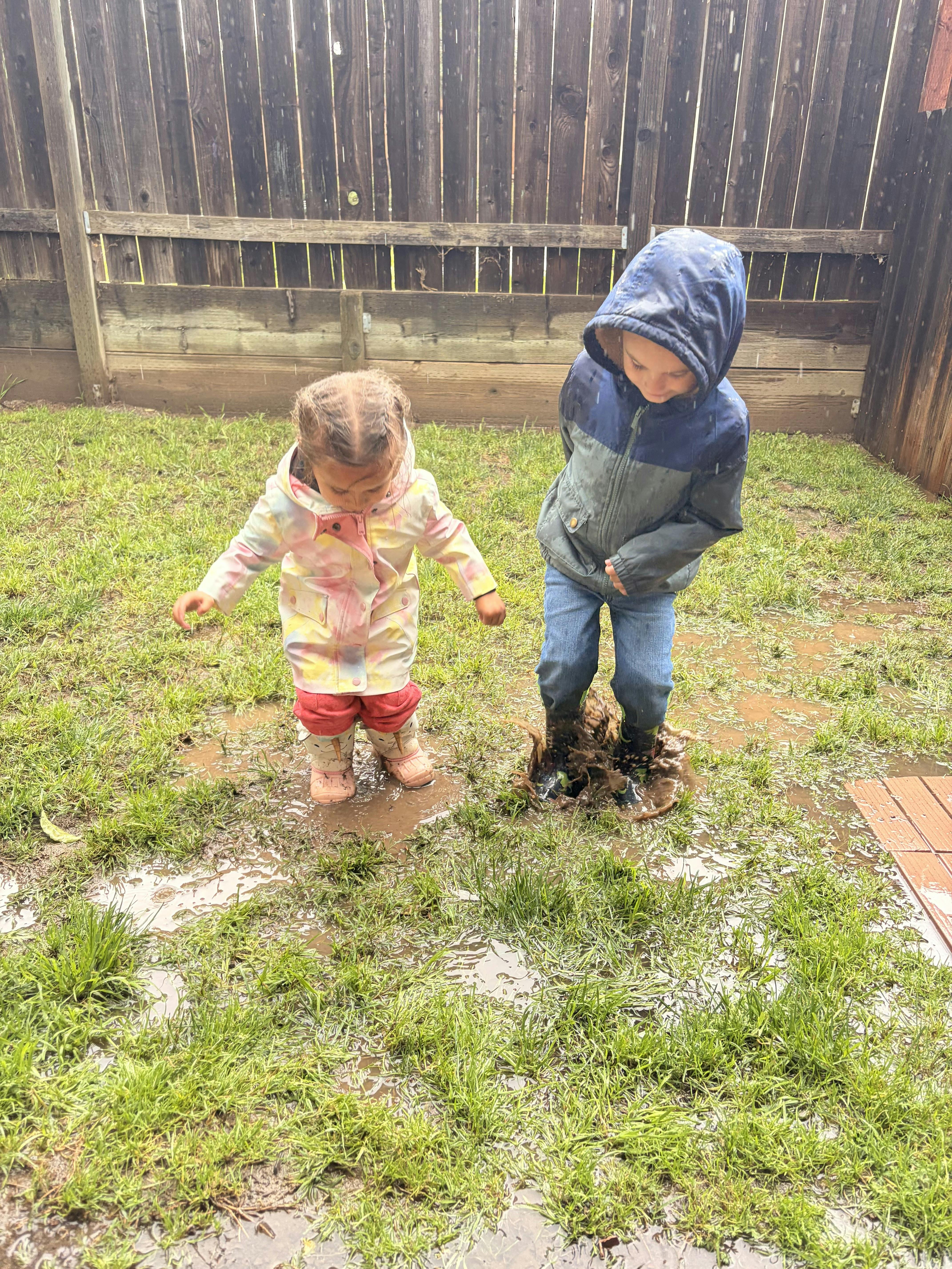 Two children wearing rain gear splashing in a muddy puddle on the grass